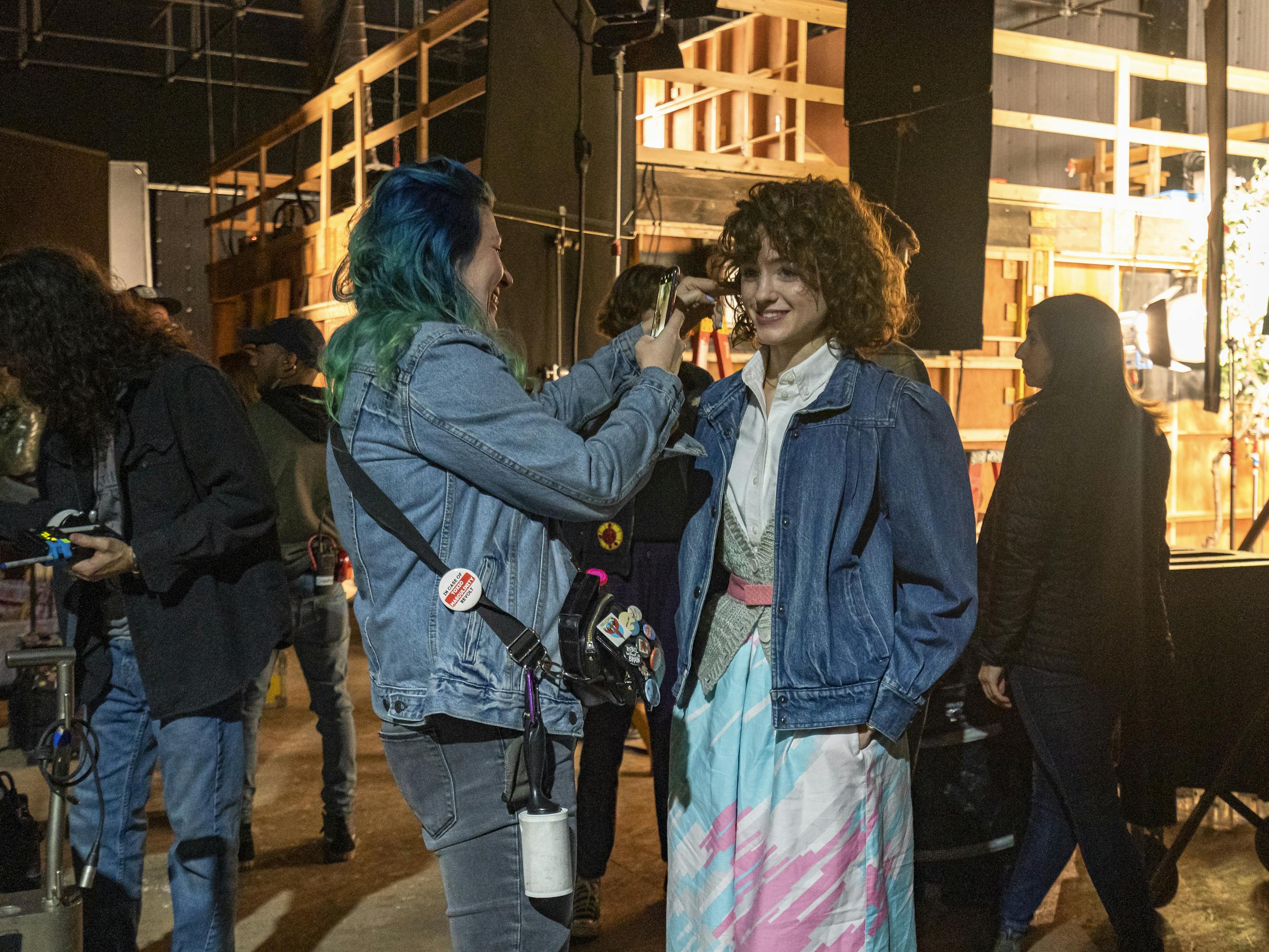 Nancy Wheeler (Natalia Dyer) wears a long skirt, denim jacket and smiles as someone adjusts her hair.