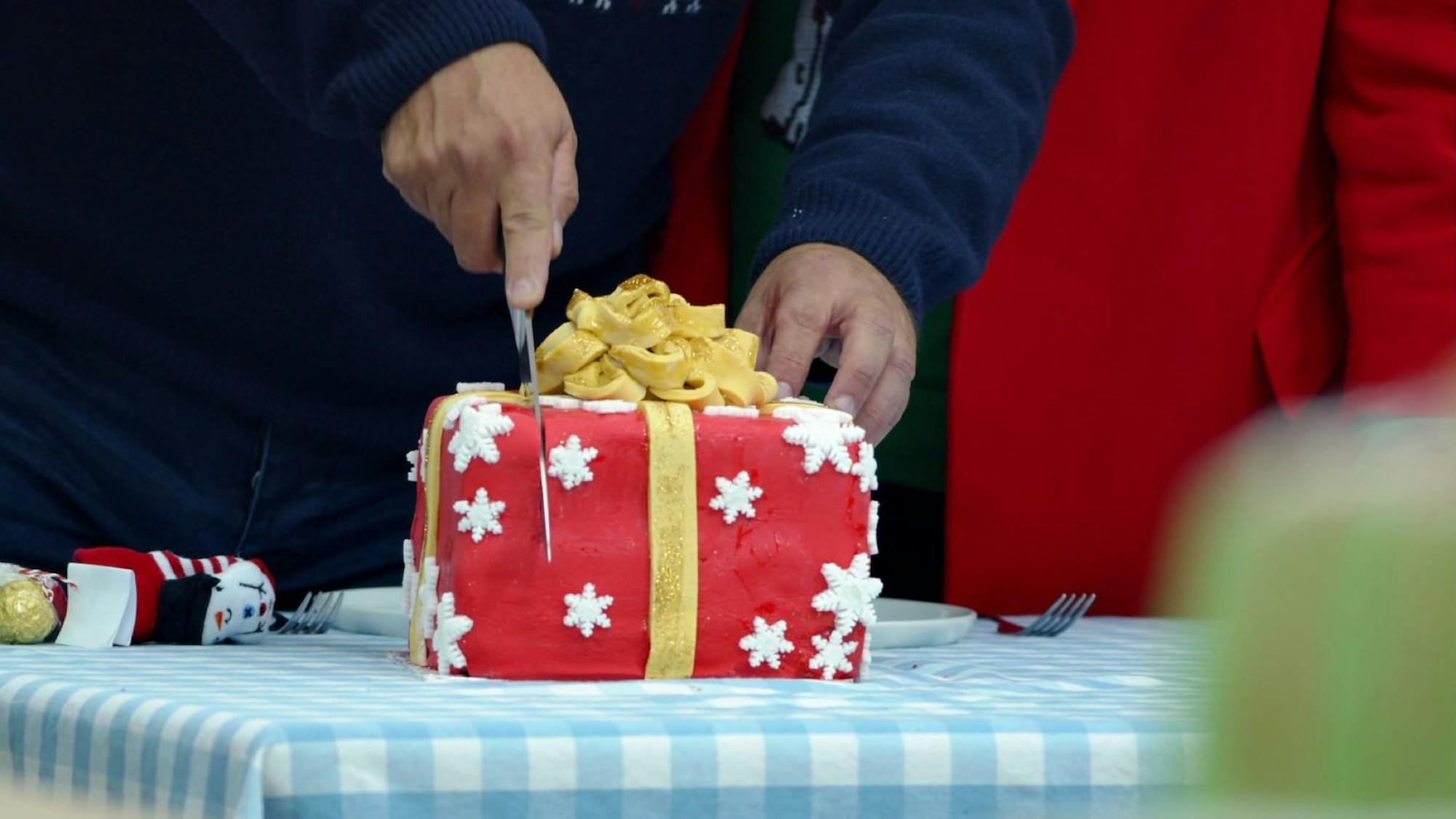 Someone cuts into a cake shaped as a present. Atop the cake is a yellow ribbon and small white stars. It sits on a blue-and-white checkered tablecloth.