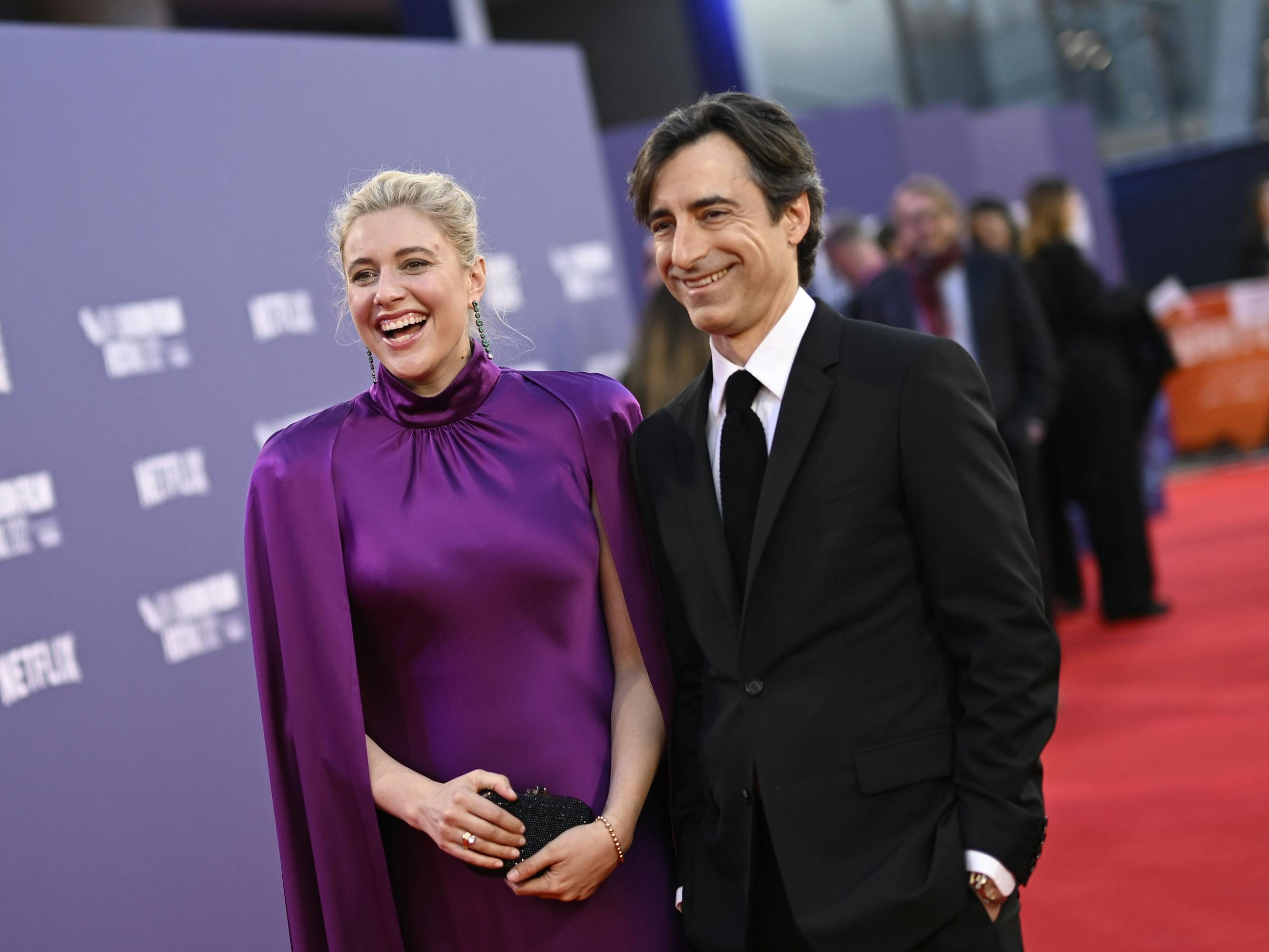 Greta Gerwig and Noah Baumbach stand on the red carpet. Gerwig wears a purple dress and holds a black clutch and Baumbach wears a dark suit and white shirt.