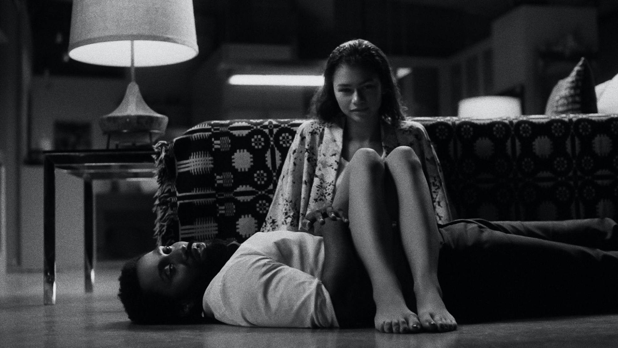 A still from Malcolm & Marie shows John David Washington, as Malcolm, lying on the floor while Zendaya, as Marie, sits with her legs swung over his torso.