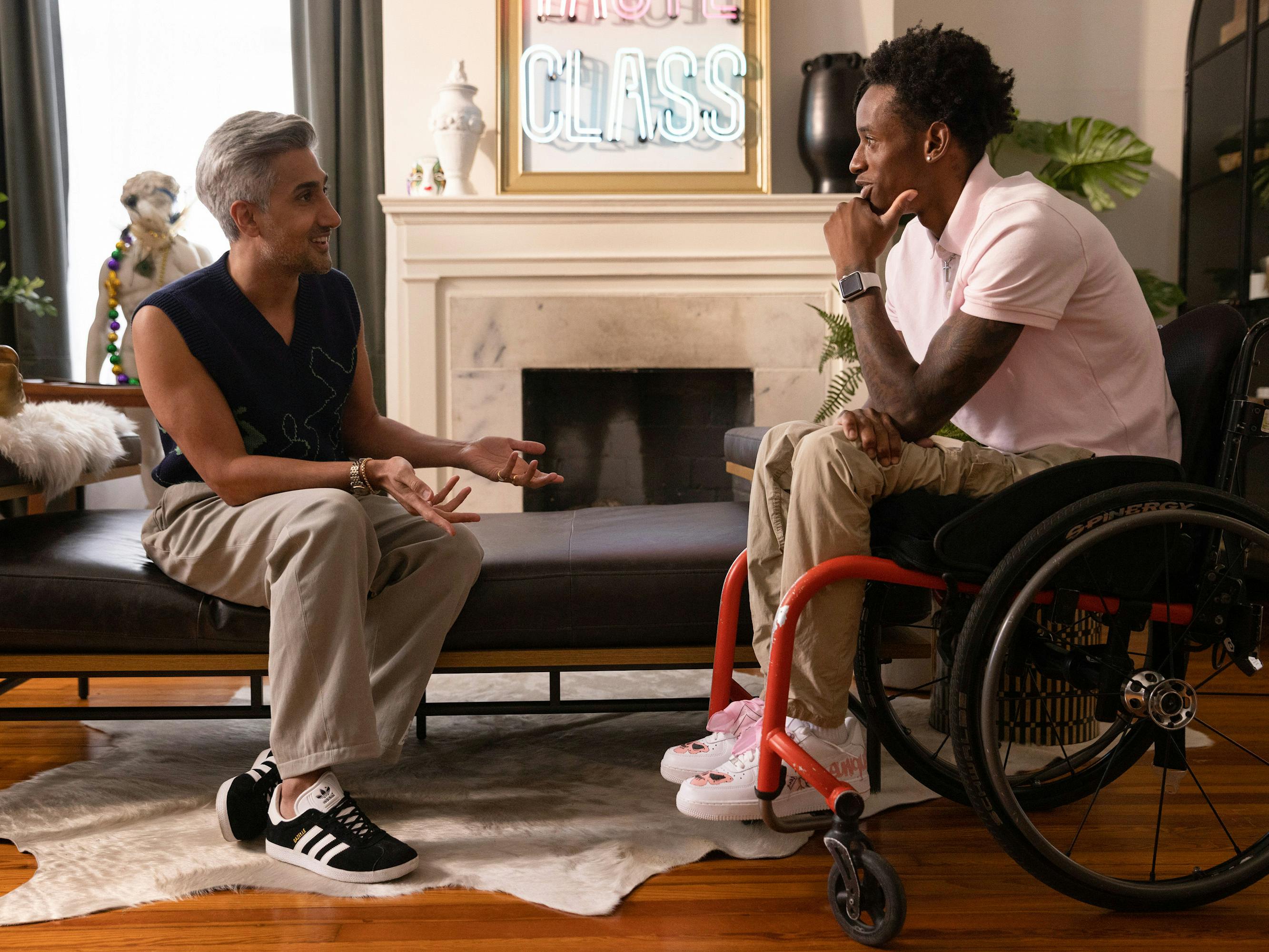 Tan France and Ray Walker (Speedy) face off in a nicely-decorated living room. Tan wears black-and-white sneakers, khakis, and a black sweater vest. Speedy wears a pink collared shirt, khakis, and sneakers, and sits in a wheelchair.