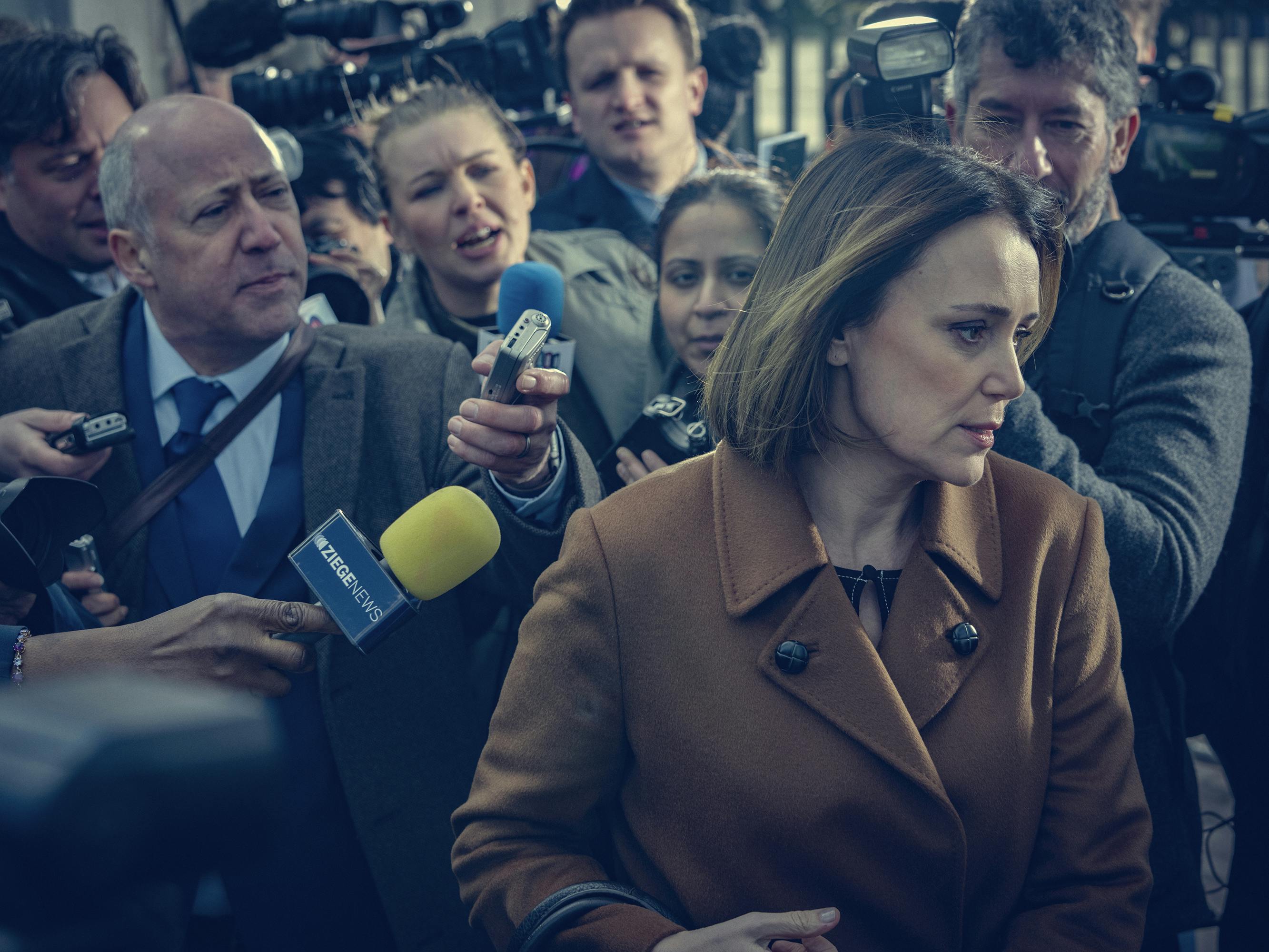 Amanda Thirsk (Keeley Hawes) wades through reporters and wears a brown trenchcoat.