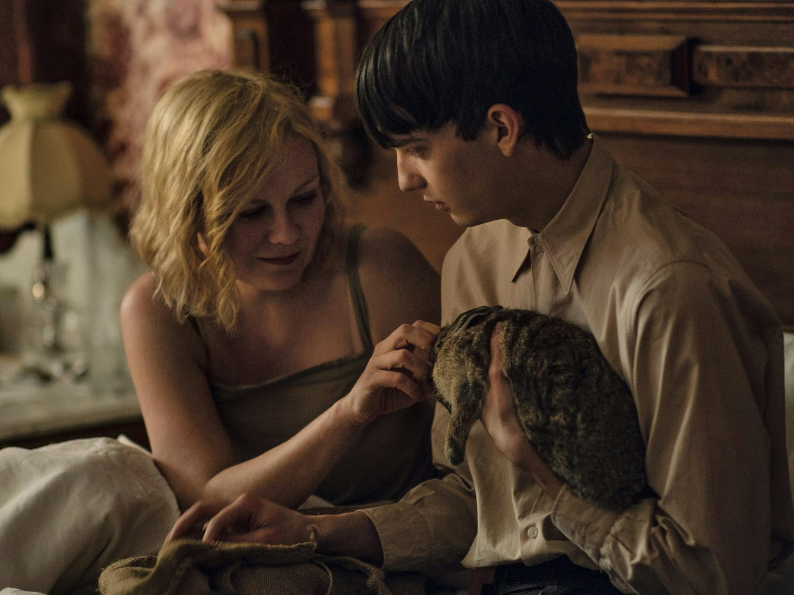 Rose (Kirsten Dunst) and Peter (Kodi Smit-McPhee) sit in bed. Rose wears a green dress and pets a bunny in Peter’s arms.