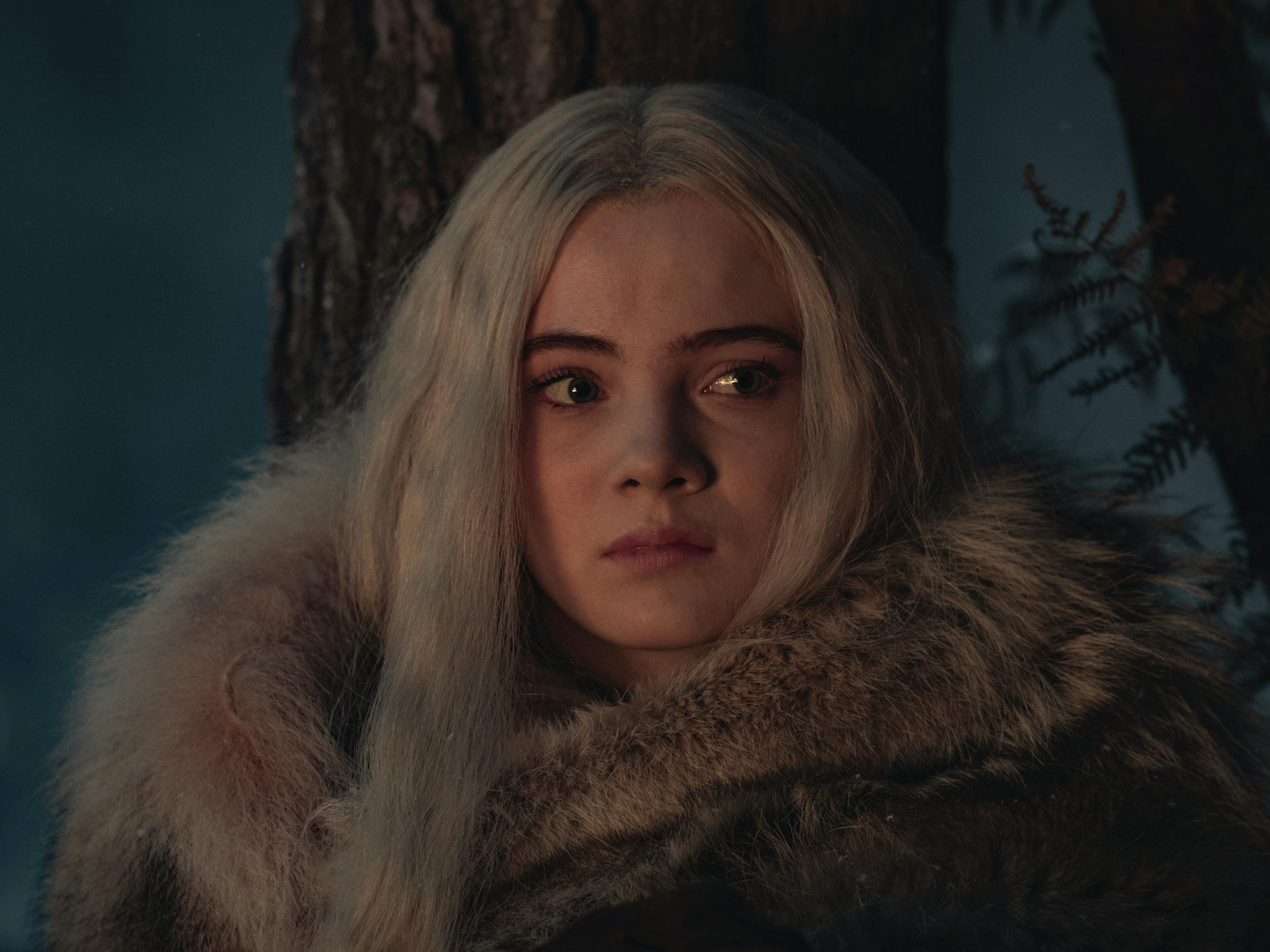 Freya Allen as Princess Ciri. She wears a fur jacket. Its nighttime and there are trees behind her. Her expression conveys concern