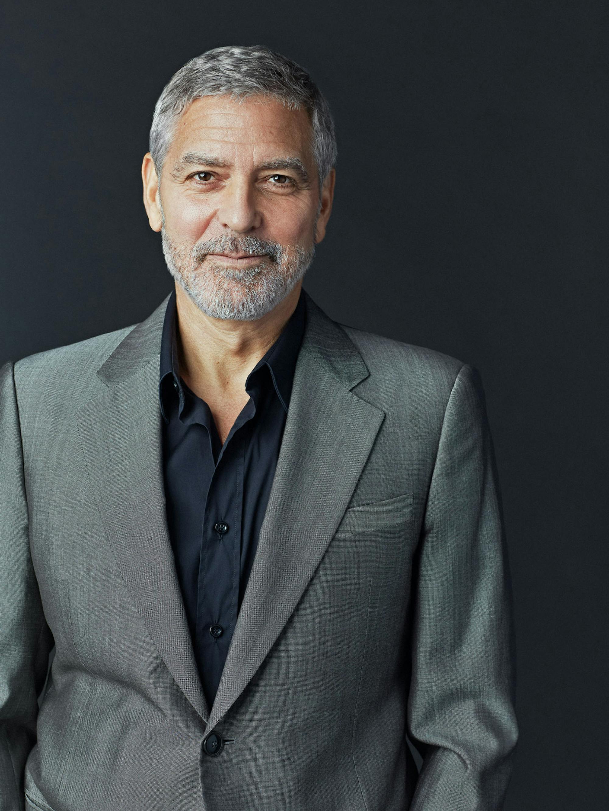 George Clooney in a gray suit against a black background. He gives you a subtle smile.