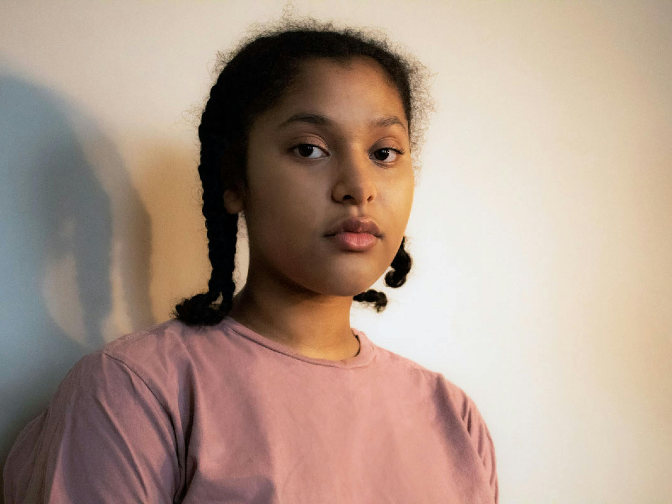 This portrait shows a girl in two short braids and a pink t-shirt. She stands against a white wall, against which her shadow falls. She looks directly into the camera, unsmiling. 