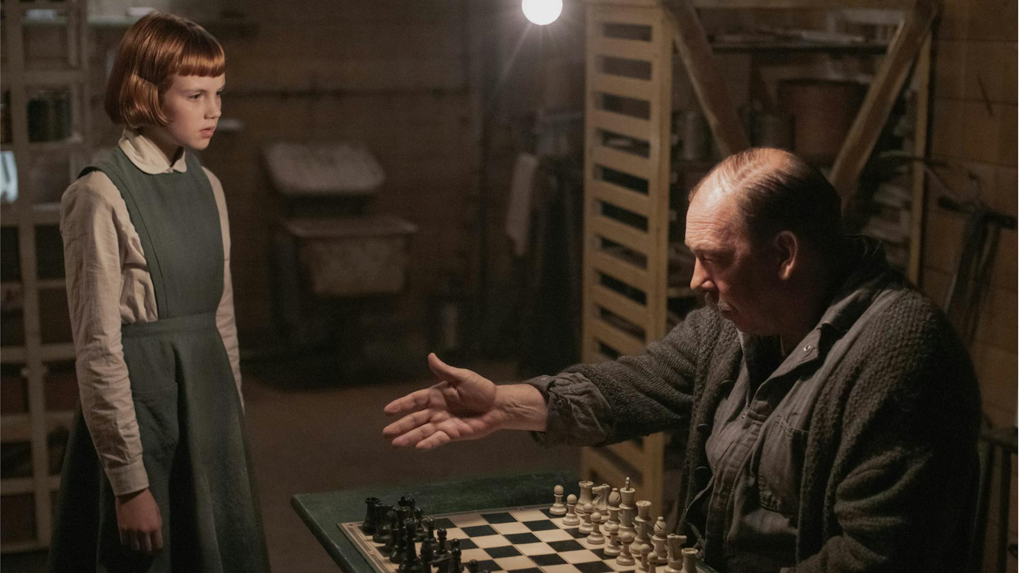 Bill Camp’s character in The Queen’s Gambit, Mr. Shaibel, gesturing for a young Beth Harmon to join him at the chess board. She wears a gray tunic, a long-sleeved buttonned down shirt, and has her signature red bob. He wears all gray, and is surrounded by his janitorial tools.