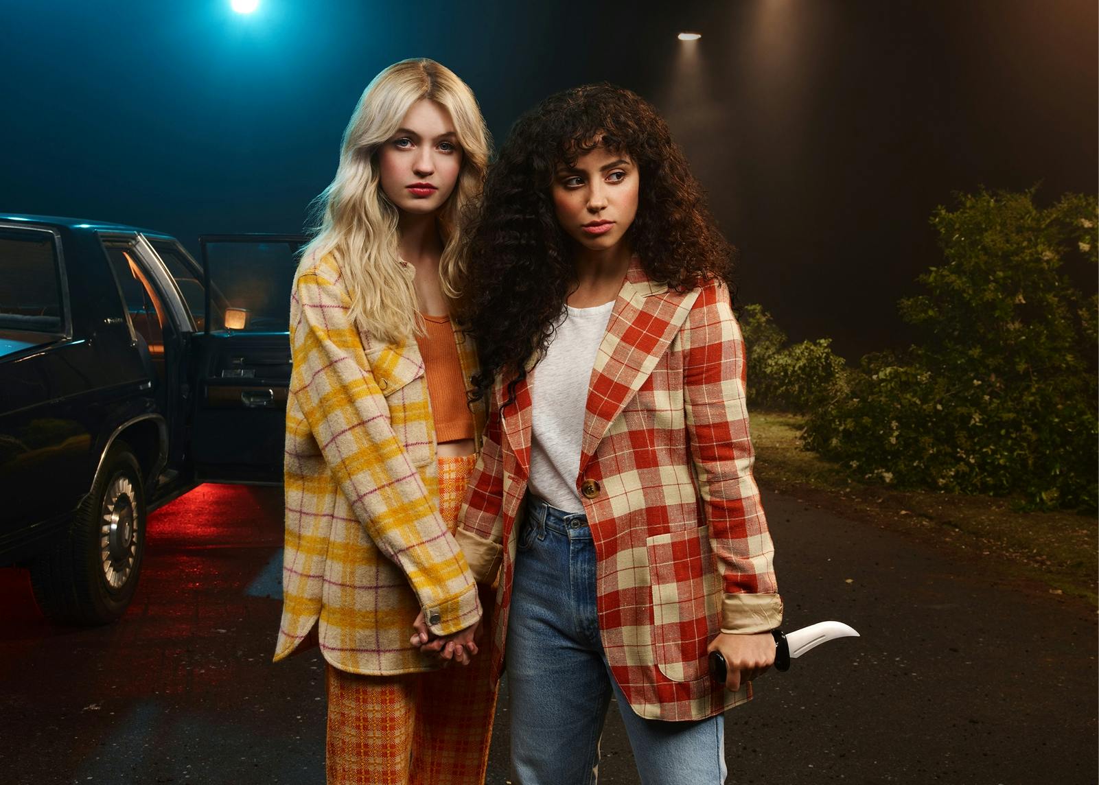 Sam Fraser (Olivia Scott Welch) and Deena (Kiana Madeira) stand holding hands in a dark, streetlight-lit shot. They both wear plaid jackets, and Deena carries a knife.
