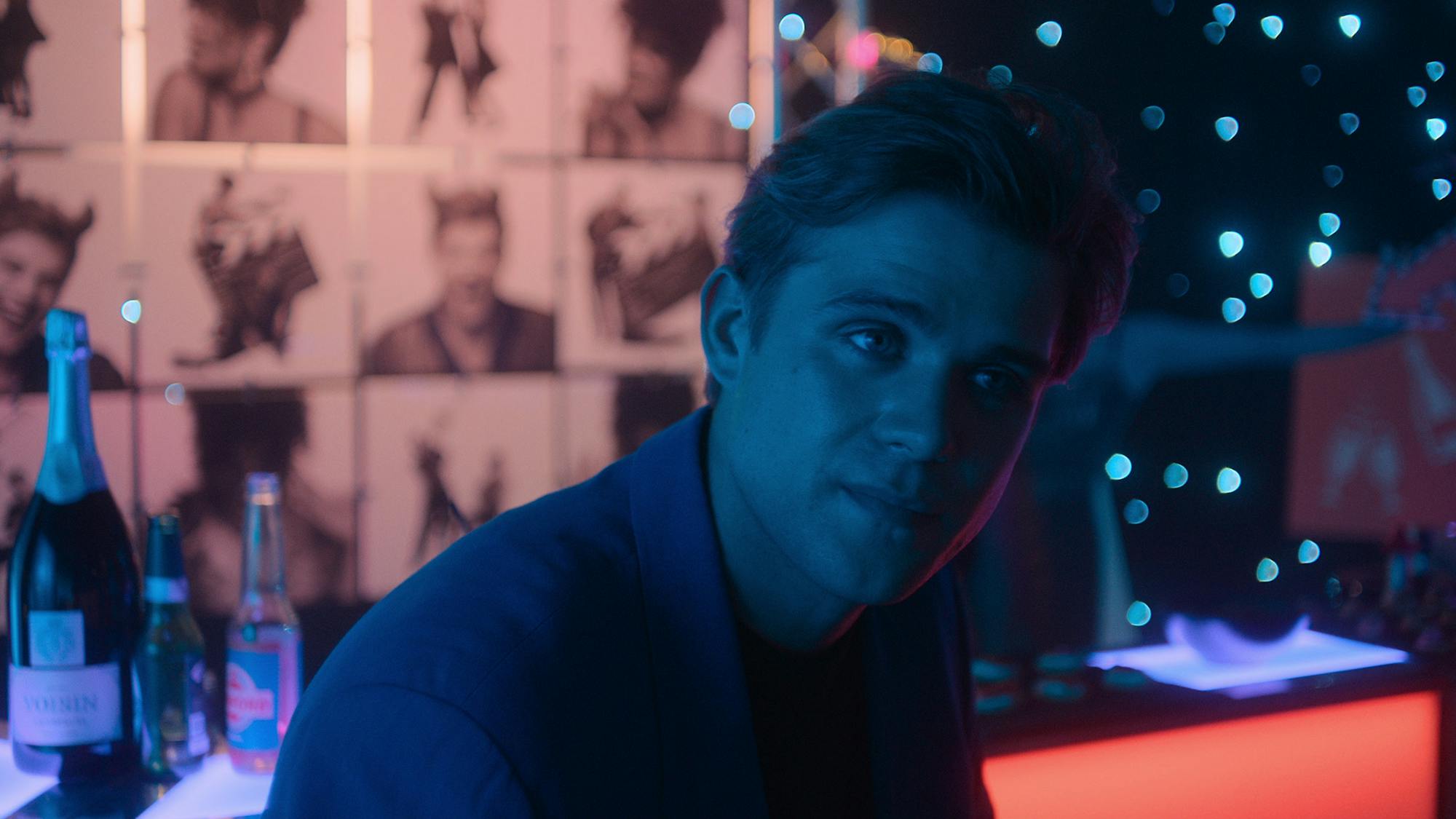 Dexter Mayhew (Leo Woodall) sits in a dark club, with drinks and pictures of himself in the background