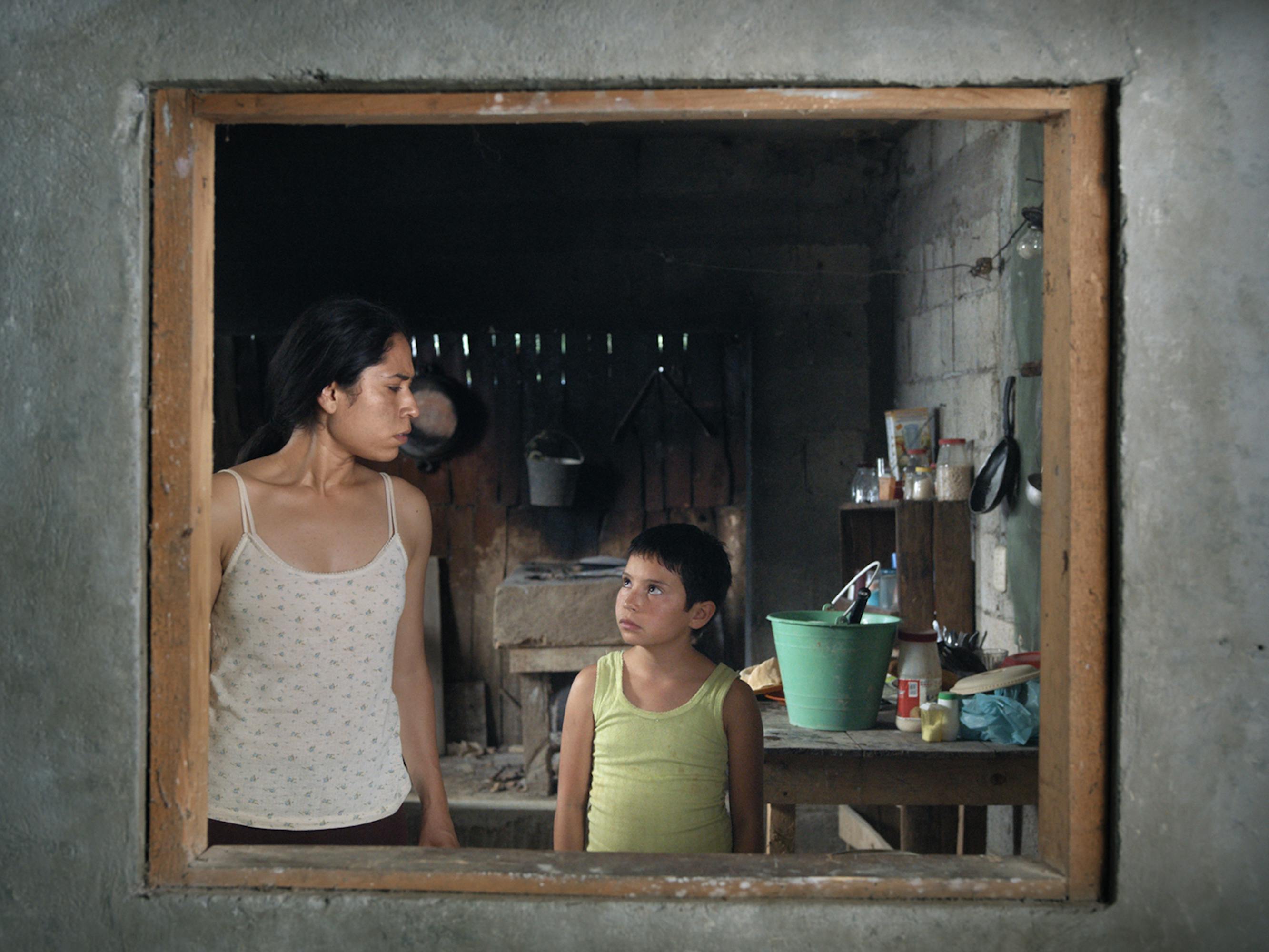 Two characters from Noche de fuego stand behind a window of a cement wall. 