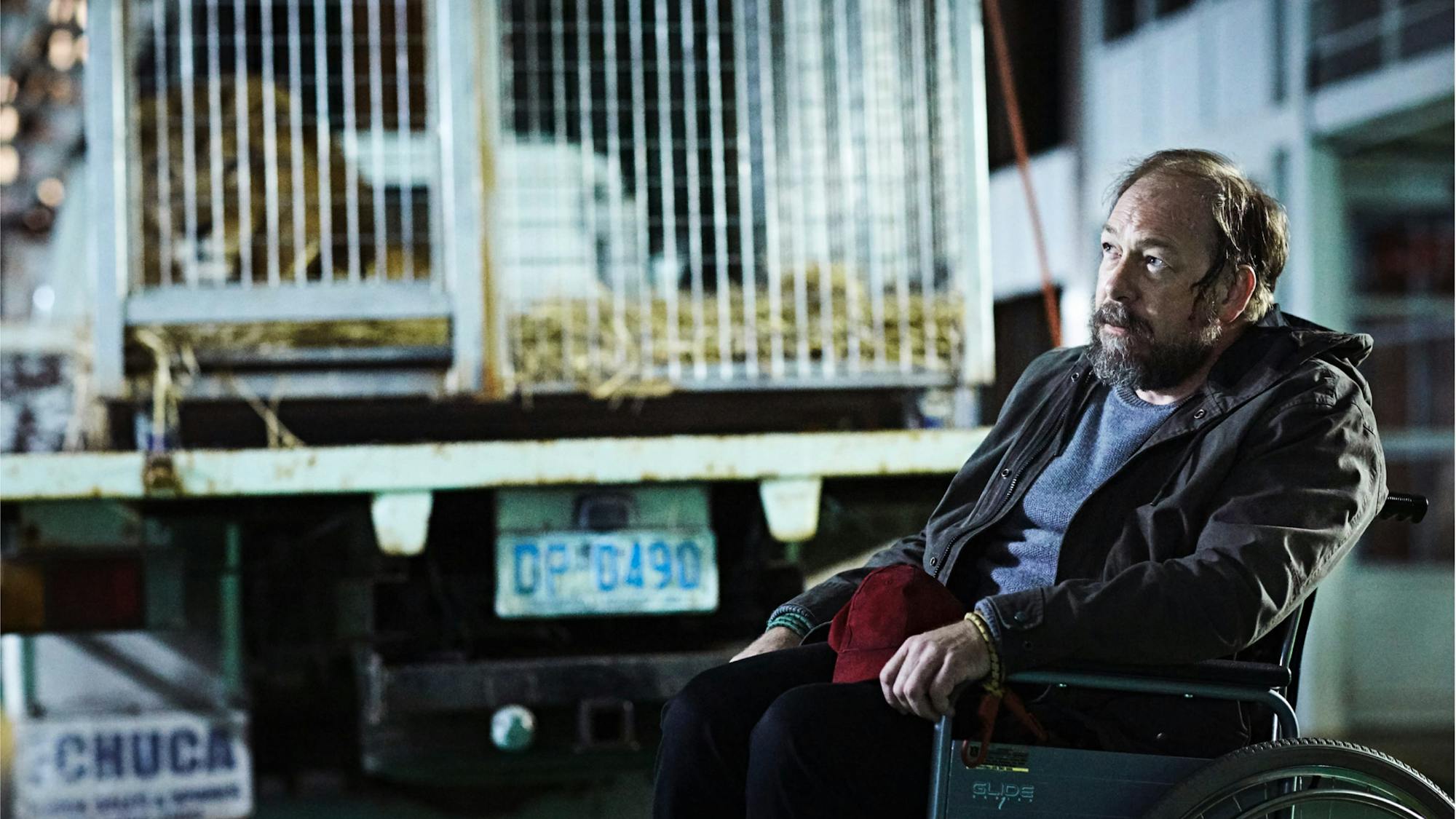 Bill Camp’s character in The Leftovers, David Burton. He wears dark clothing and a blue shirt, and sits in a wheelchair. There is some kind of truck in the background with a lisence plate that reads DP 6490.