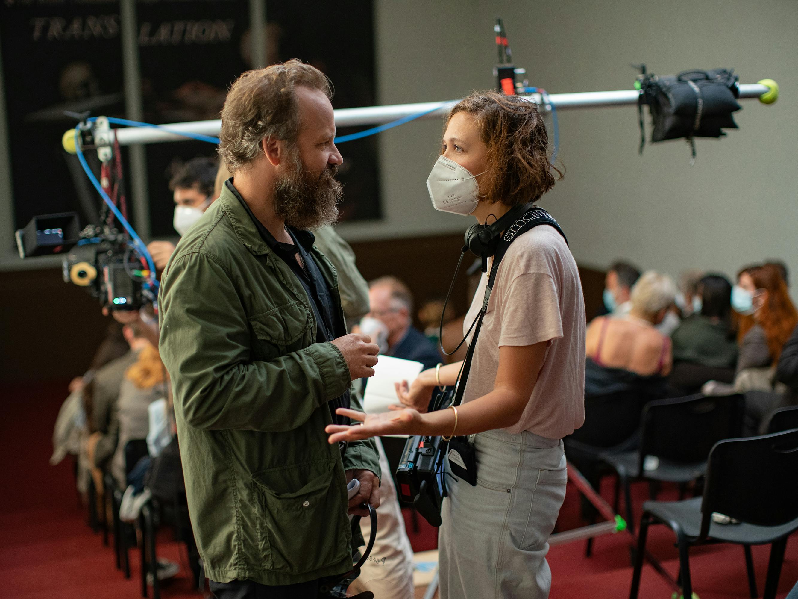 Peter Sarsgaard and Maggie Gyllenhaal stand among equipment and black plastic chairs. Sarsgaard wears a green jacket and Gyllenhaal wears khakis and a pink shirt.  