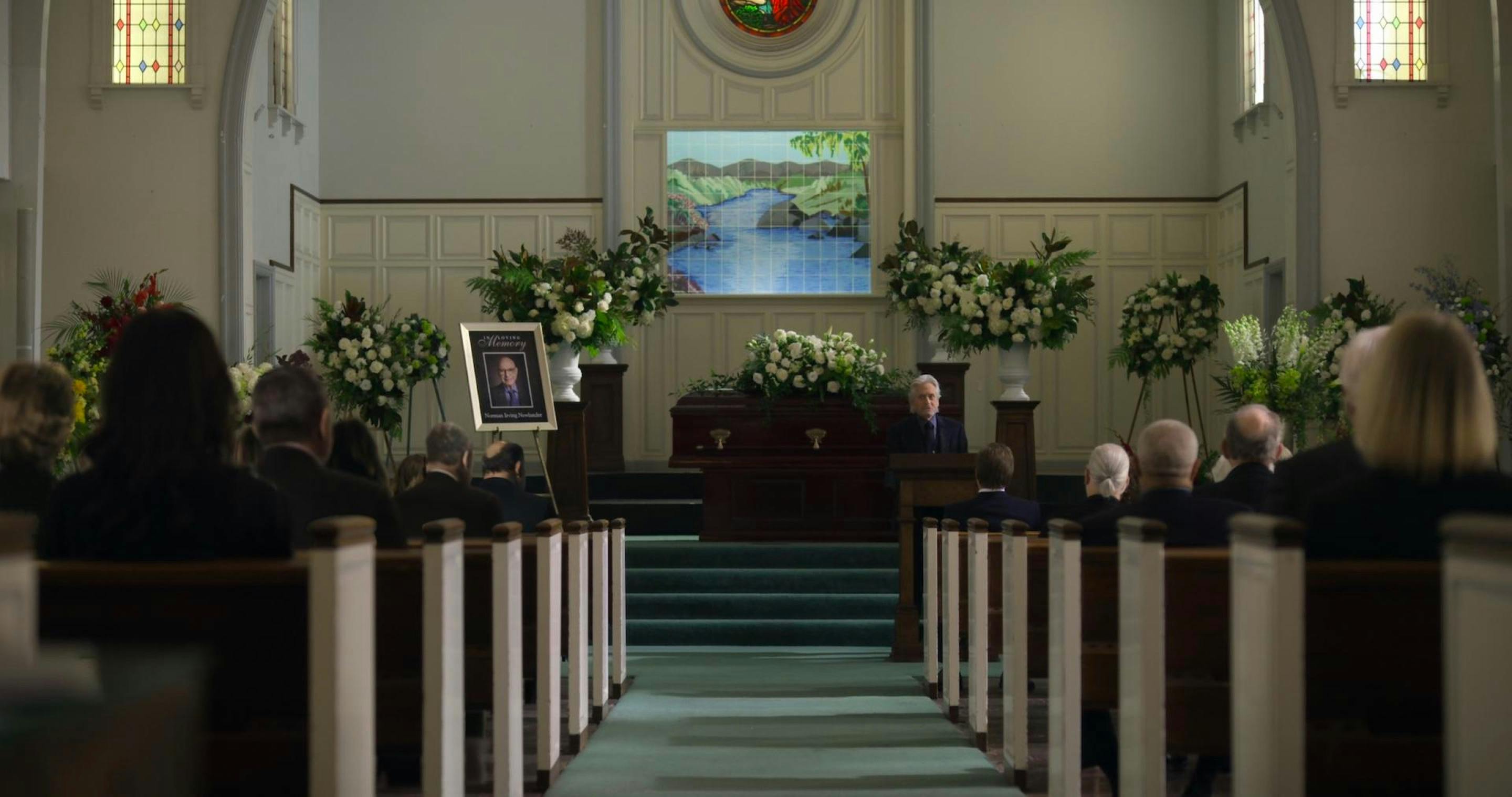 This shot shows a brightly lit funeral. Pews line either side, and we see the backs of many people’s heads. Michael Douglas’s character, Sandy Kominksy, delivers an unexpected eulogy for his best friend, Norman Newlander (Alan Arkin). This is a poster with an image of Newlander to the left of the coffin, and flowers line the back wall.