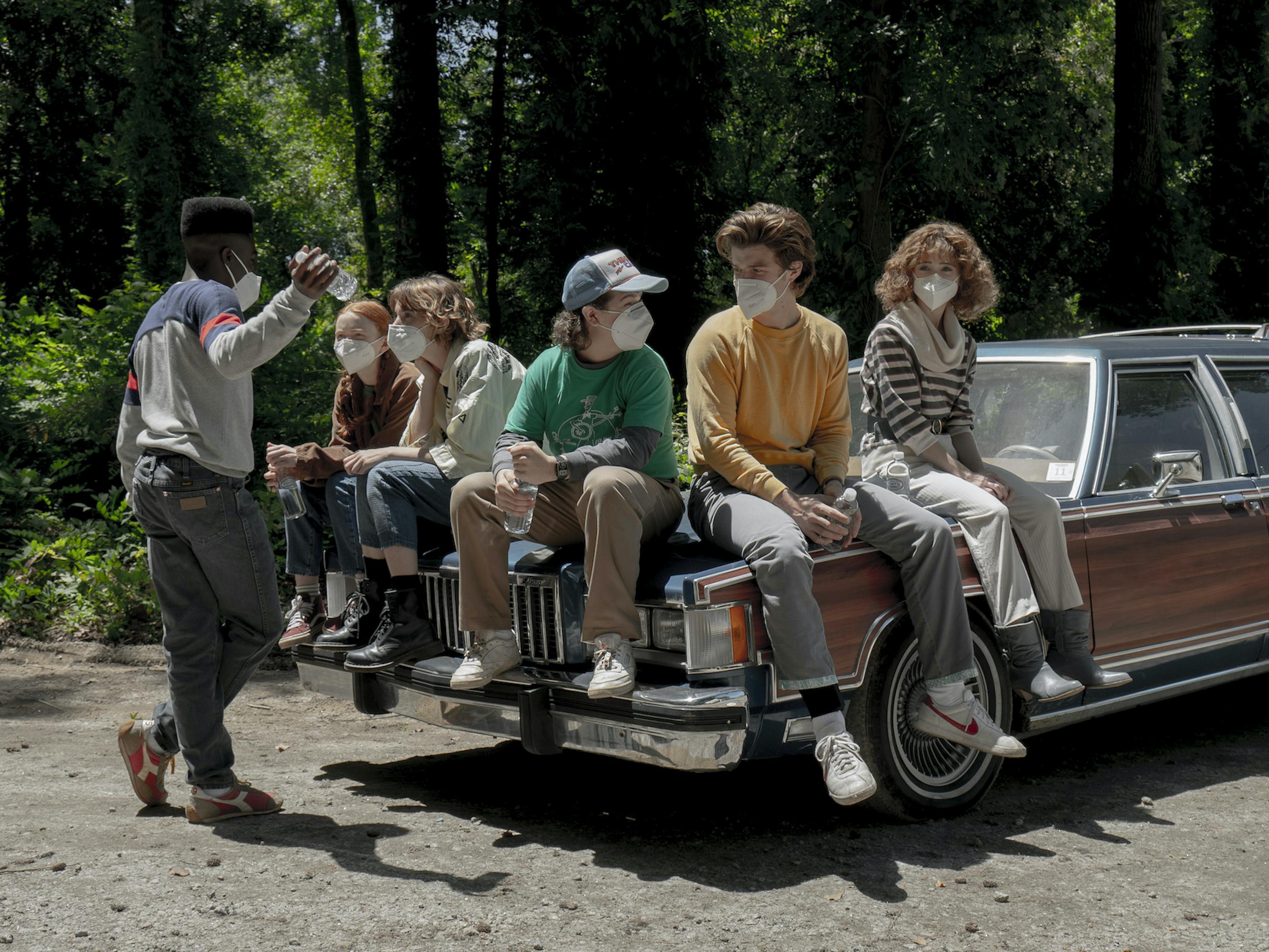 The cast of Stranger Things sit on a station wagon wearing masks in an outdoor sunny scene.