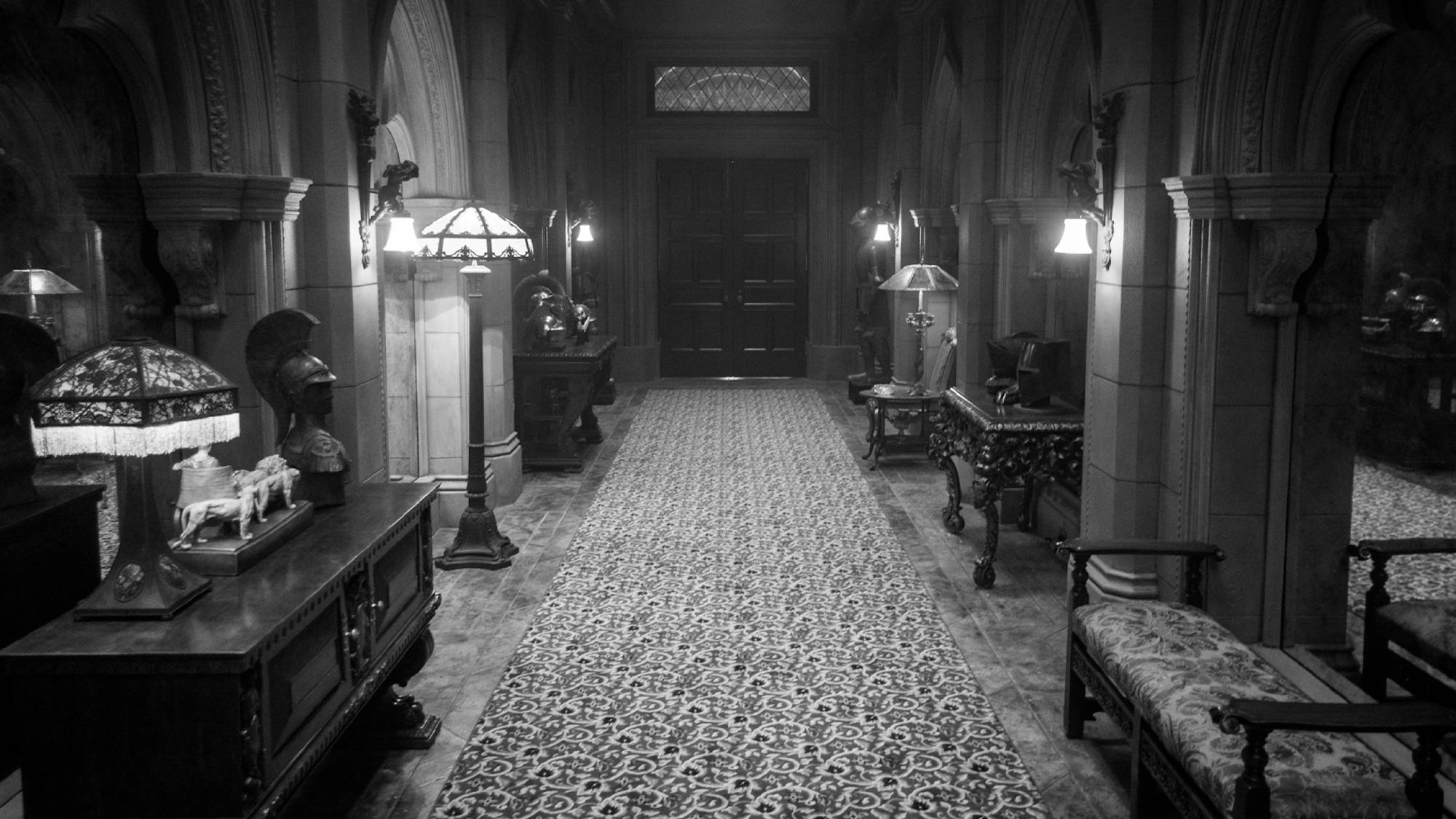 A Hearst Castle interior set includes a long hallway featuring an equally long, intricately patterned runner. The passage is built from large stones and ends in a set of double doors. The sides of the hall are lined with dark wood tables, upholstered benches, and an assortment of table, standing, and mounted lamps. 
