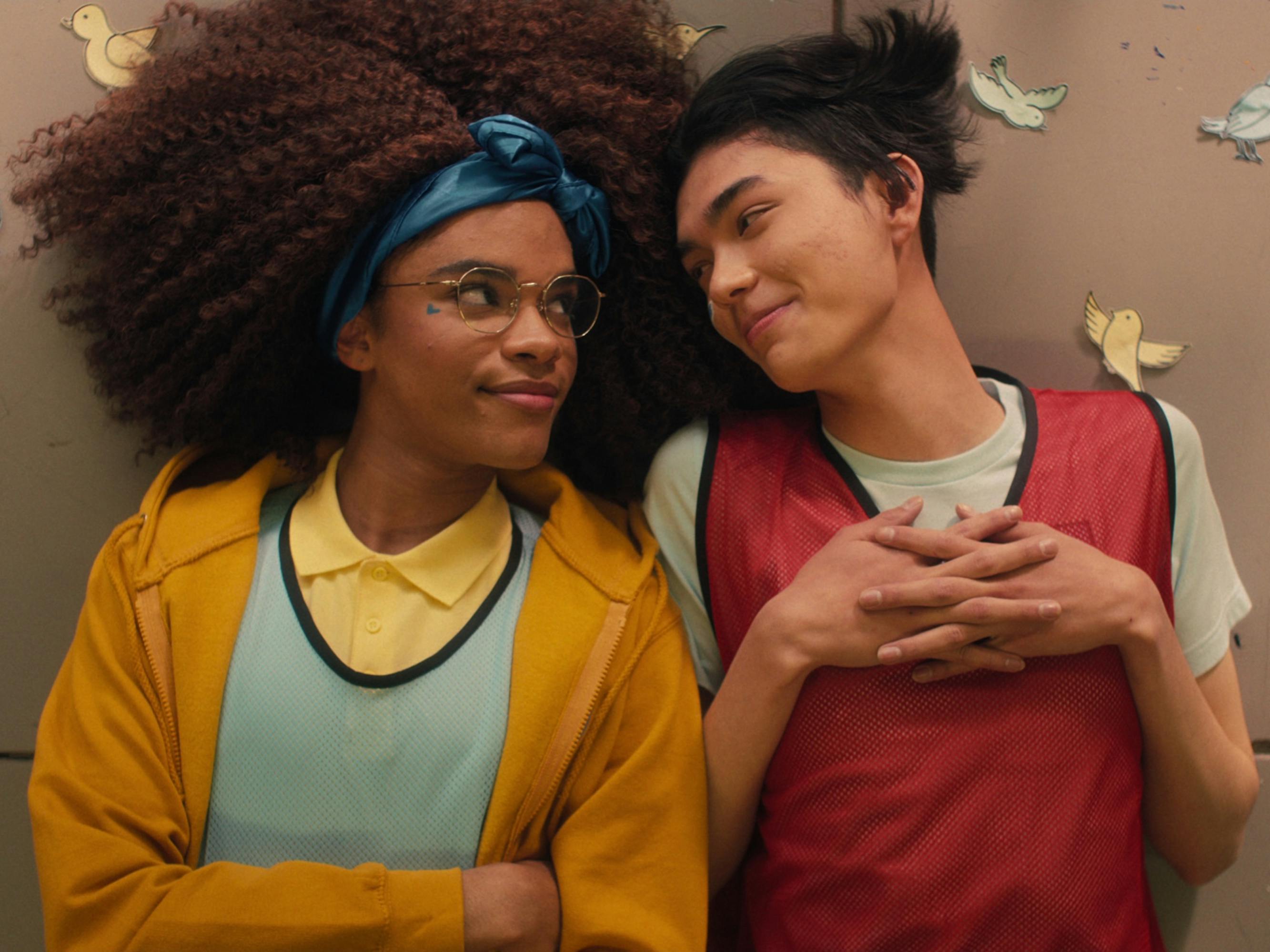 Elle (Yasmin Finney) and Tao Xu (William Gao) lie on the ground and look at each other lovingly. She wears a yellow hoodie and blue headband and he wears a red pinny.
