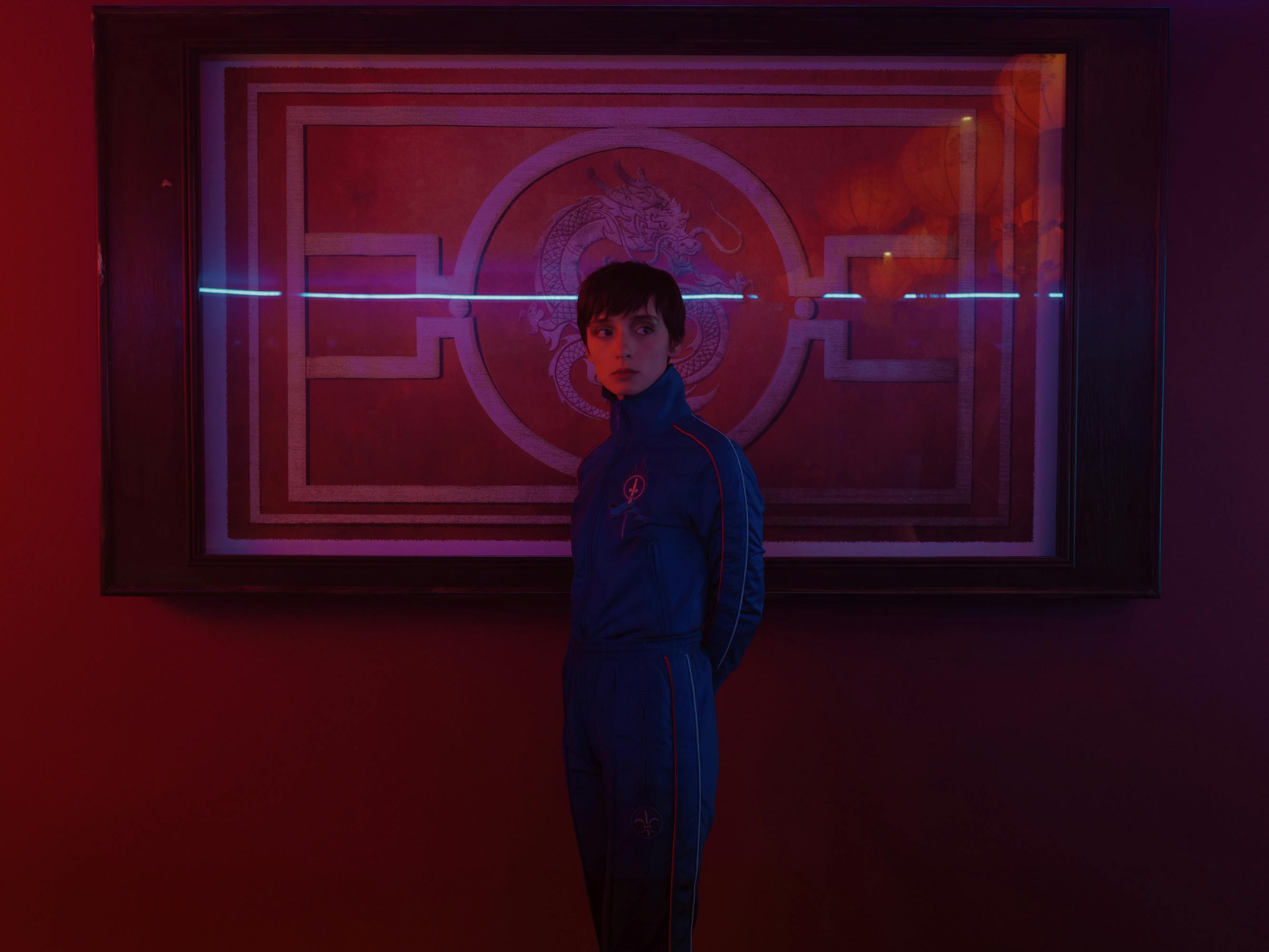Miu (Angela Bundalovic) wears a blue suit in a red-lit scene.