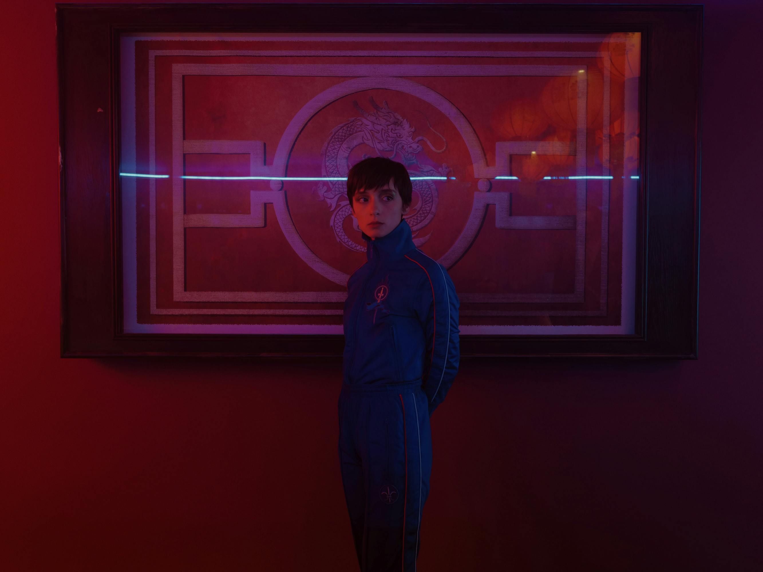 Miu (Angela Bundalovic) wears a blue suit in a red-lit scene. 