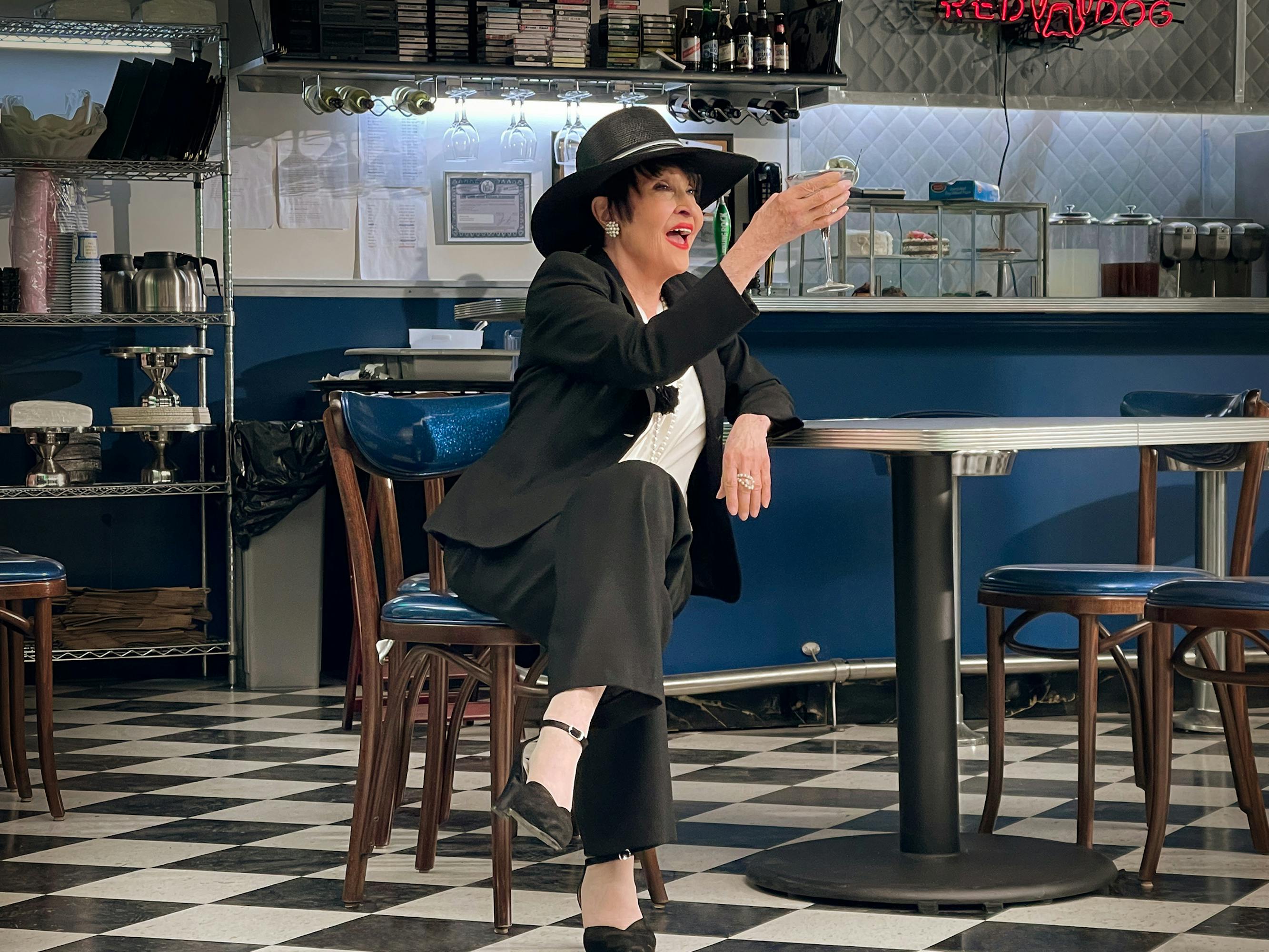 Chita Rivera wears a black suit and wide brimmed hat as she sits in the Moondance Diner.