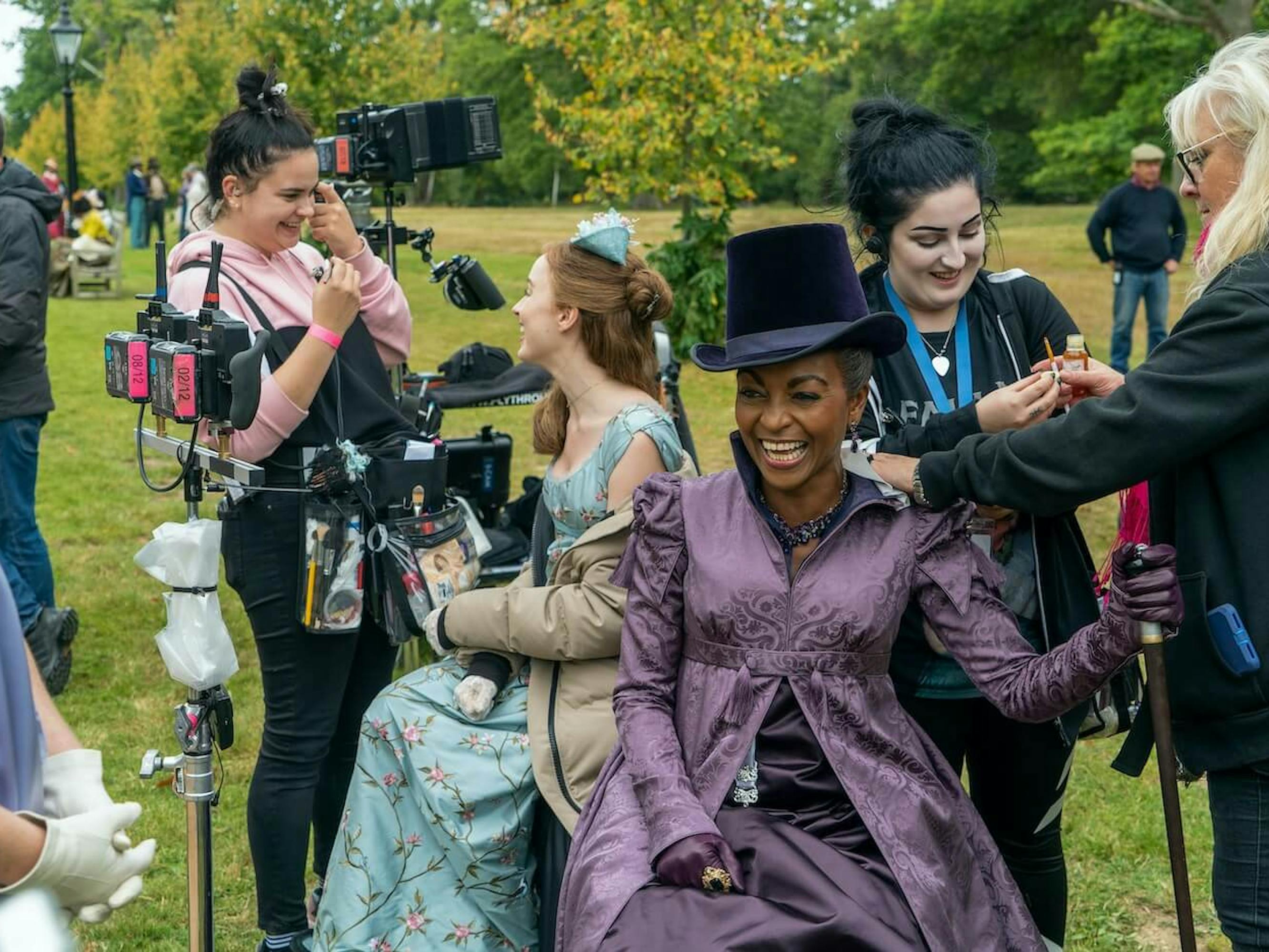 A behind-the-scenes shot of Daphne Bridgerton (Phoebe Dynevor), Lady Danbury (Adjoa Andoh), and the Bridgerton crew. Dynevor and Andoh wear full costume, including a light blue hair accessory, and purple velvet hat, respectively. The crew touch them up, and they all smile and laugh. The happy scene takes place in a field, surrounded by green and yellow trees, and camera equipment.