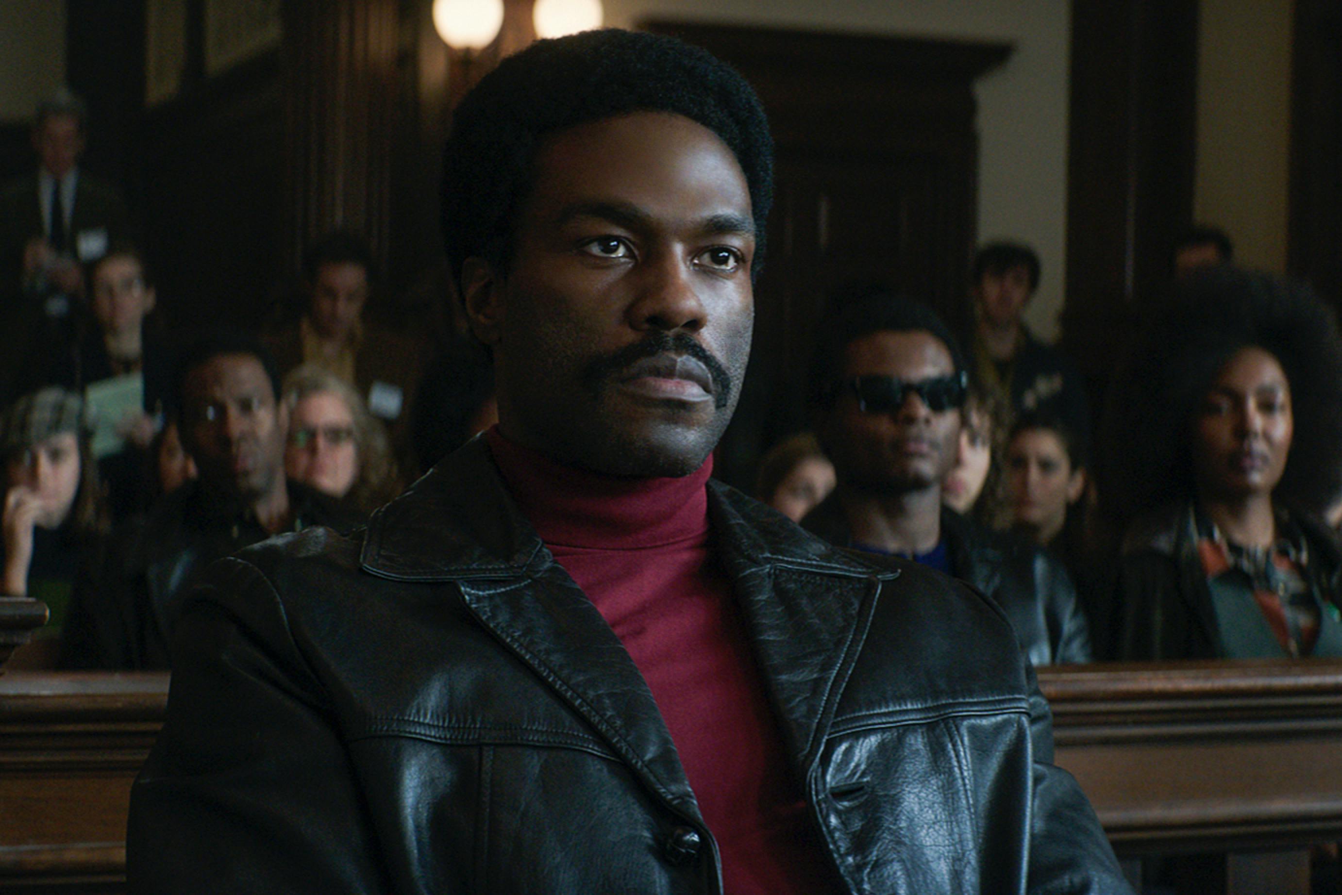 Bobby Seale, played by Yahya Abdul-Mateen II, in a courtroom scene from The Trial of the Chicago 7. Seated as a defendant, he wears a red turtleneck and black leather jacket. He is stone-faced, but behind his eyes we can see power and righteous defiance. He looks toward the bench, which is out of view. Behind him, in the gallery of the court, are several members of the Black Panther Party. 