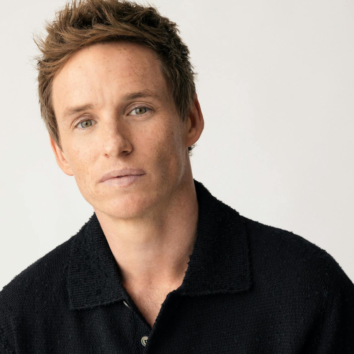 Eddie Redmayne wears a dark shirt and poses against a white background.