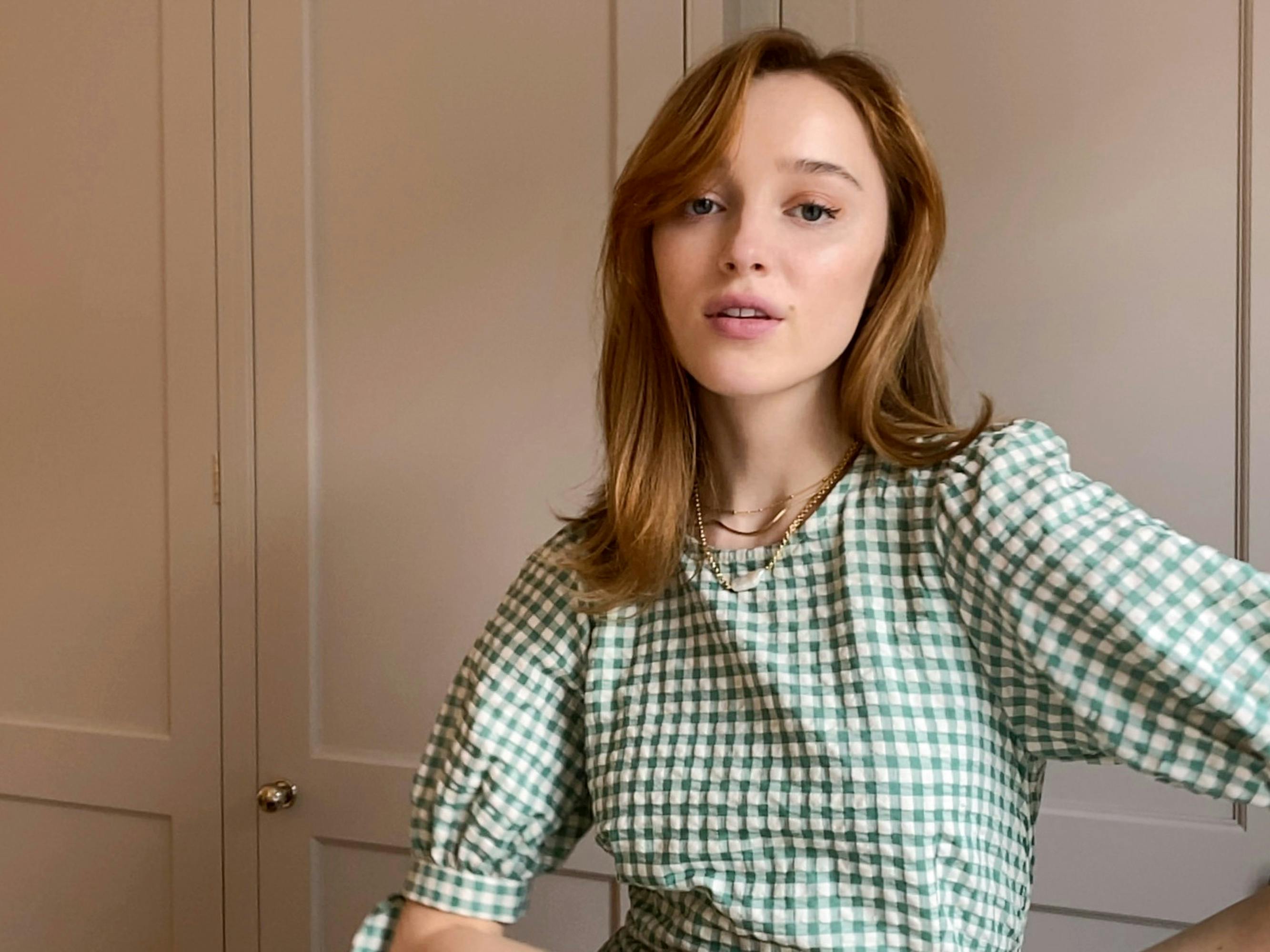 The actress wears the green gingham dress and gold necklaces. There are some cabinets in the background, and she looks directly at the camera.