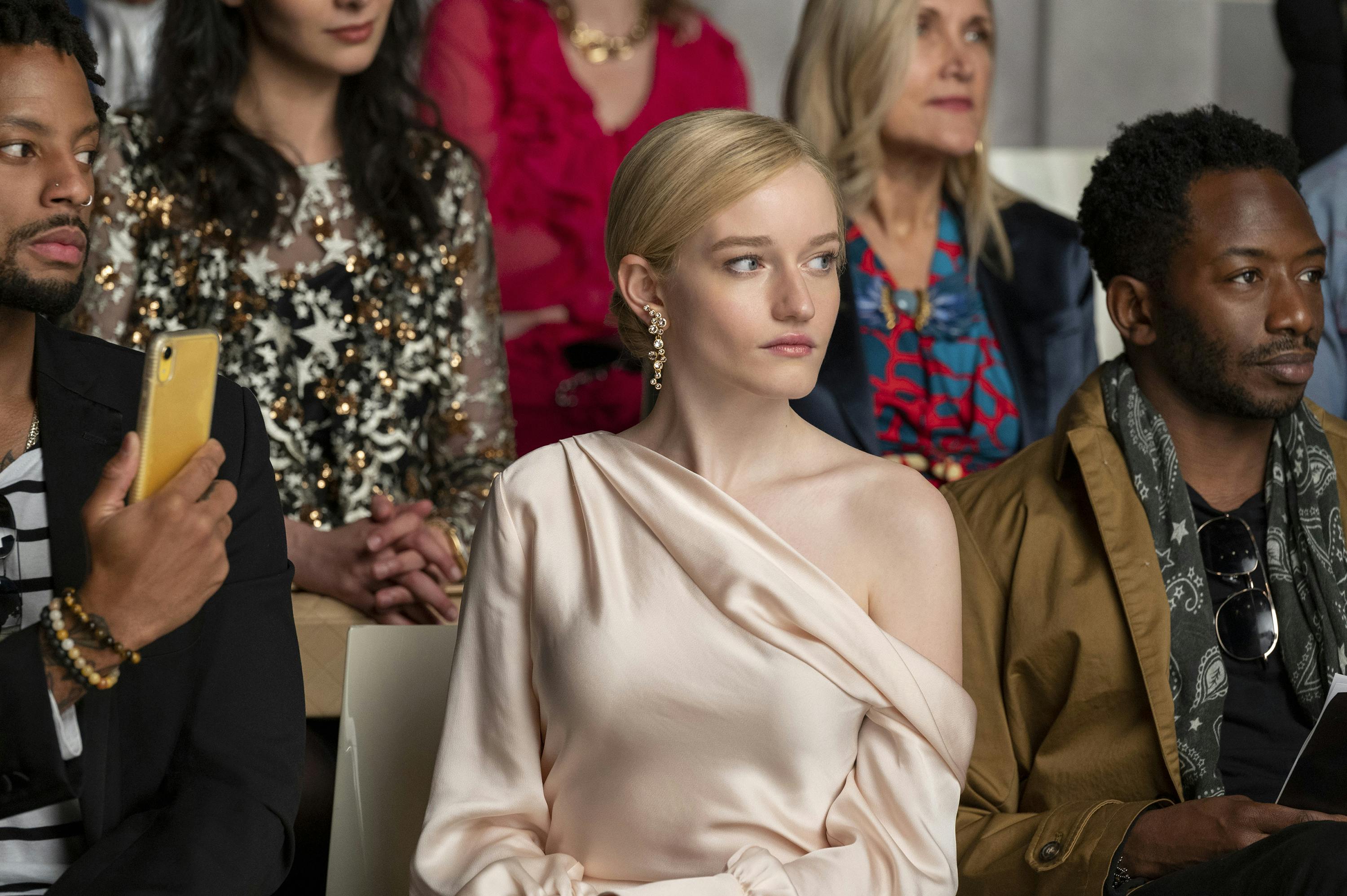 Anna Delvey wears a pale pink dress and sits front row at a fashion show.