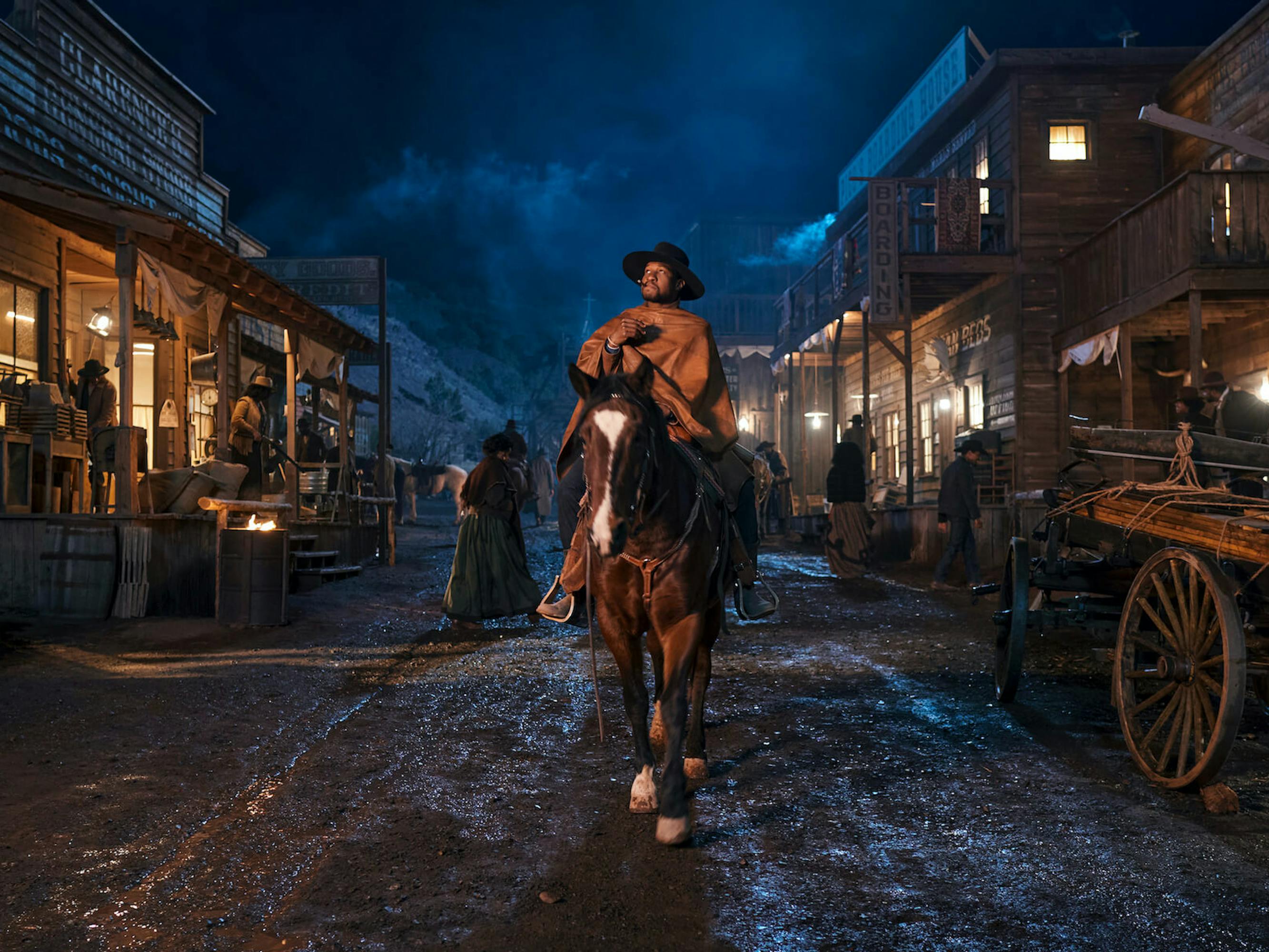 Nat Love (Jonathan Majors) rides a beautiful brown and white horse through a muddy street at nighttime. The scene is lit by house lights on either side of Majors path, and people mingle about. Majors smokes and wears a wide brimmed hat and poncho.