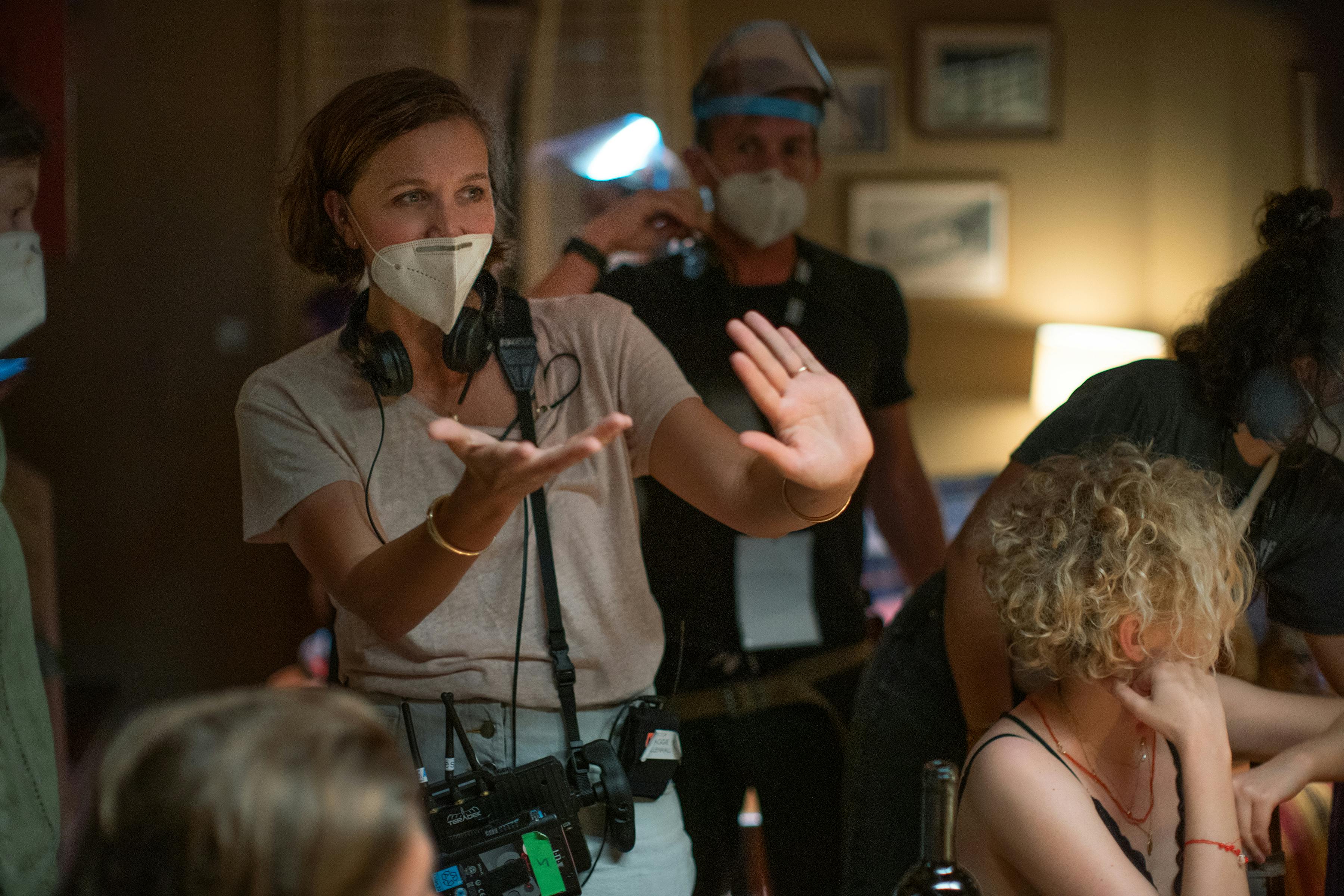 Maggie Gyllenhaal wears a white mask, tan shirt, and equipment. Other The Lost Daughter crew members mingle about.