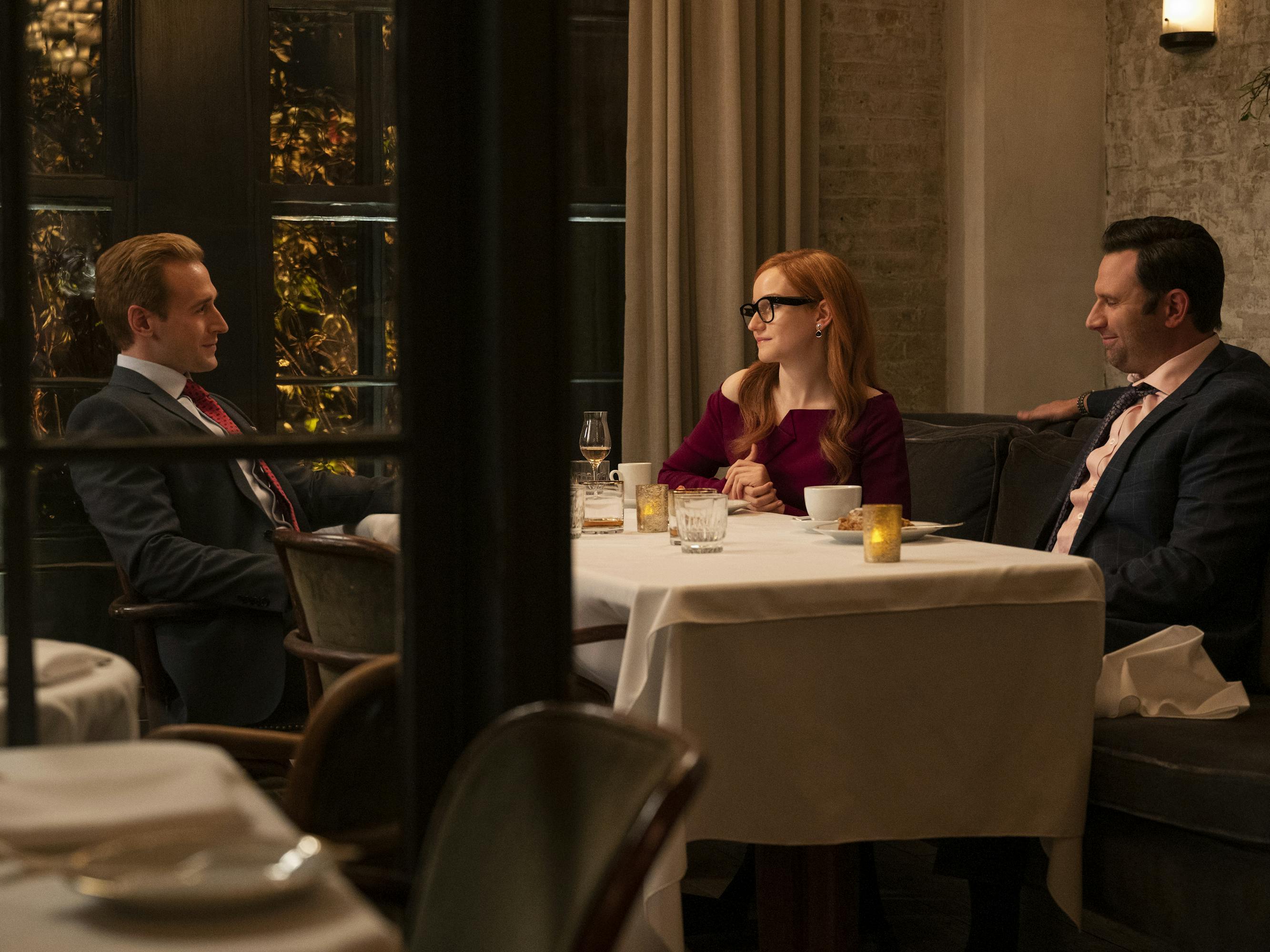 Chris Cafero, Anna Delvey, and the Jupiter Group representative sit at a dinner table. The men wear suits and Delvey wears a red dress and black framed glasses. On the table is wine, candles, and glasses of water.