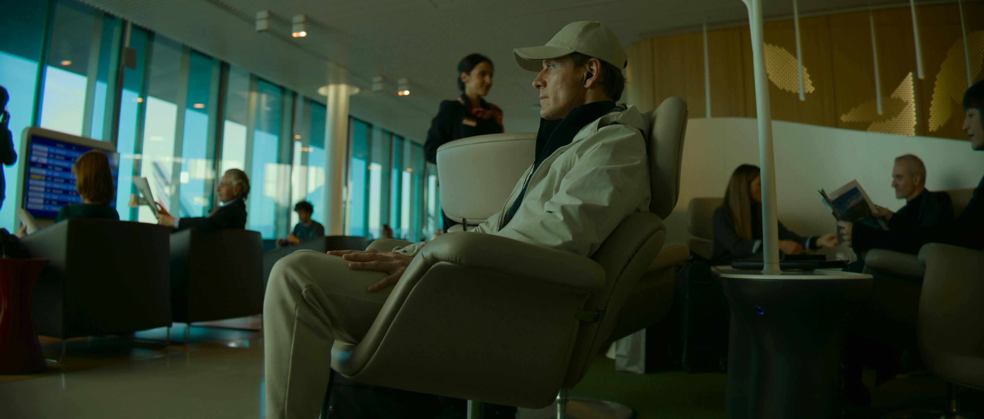 The Killer (Michael Fassbender) sits in an airport looking shady in an all-beige outfit.