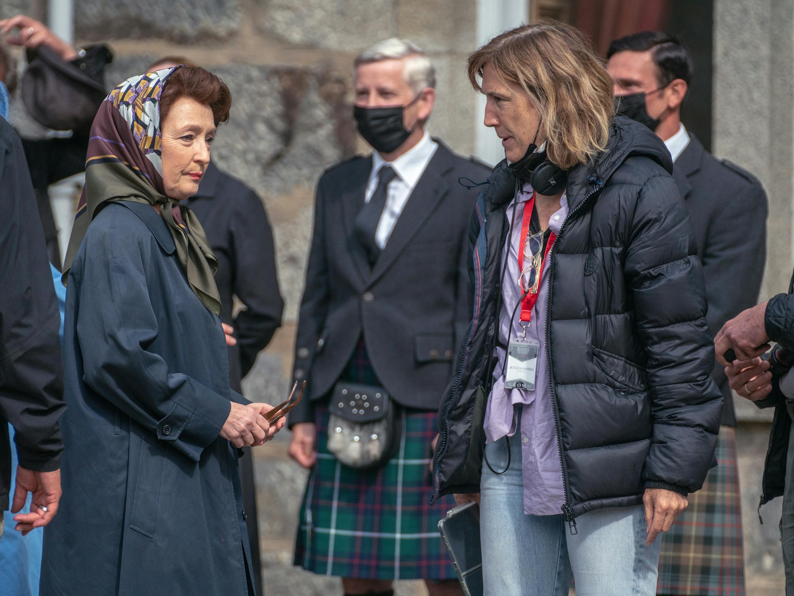 Lesley Manville behind the scenes of The Crown. She wears a navy coat and a chic headscarf. Behind her are some men in kilts. Manville talks to a woman in a black puffer jacket and jeans.