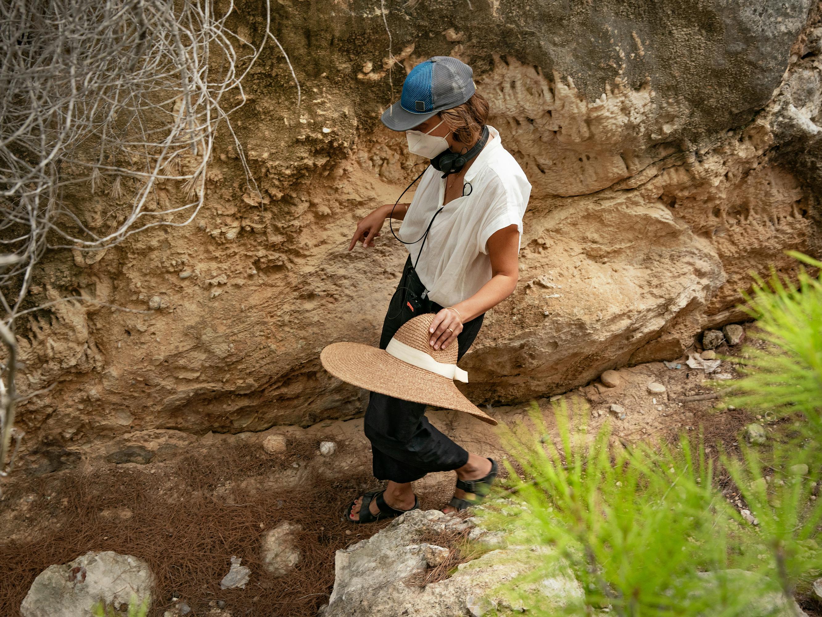 Maggie Gyllenhaal wears dark pants, dark sandals, a white shirt, a blue-and-gray hat, and carries a wide-brimmed straw hat with a white ribbon. She walks down a muddy path lined with lime green bushes.