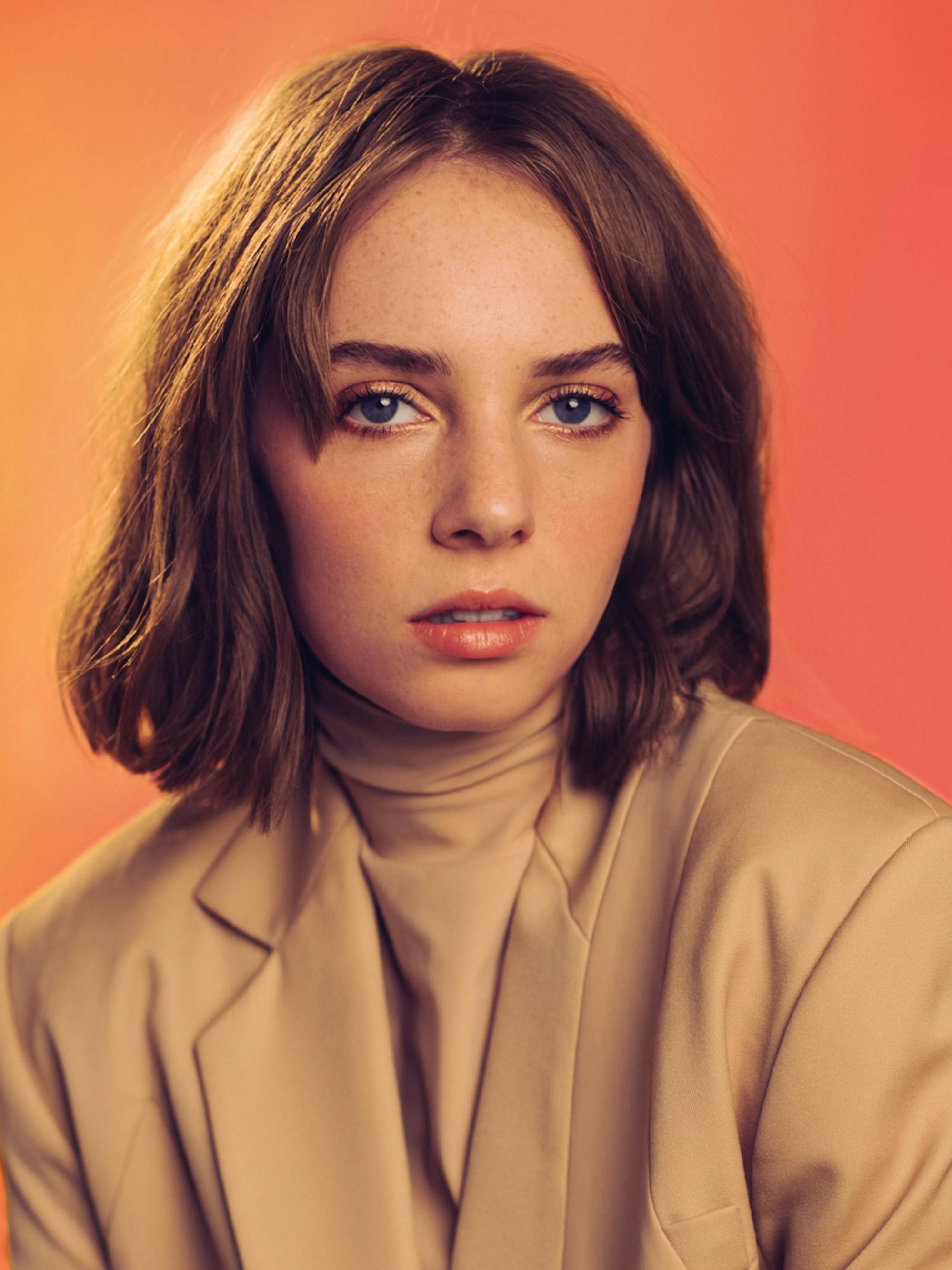A close-up of Maya Hawke of Stranger Things in front of a sunset orange background.