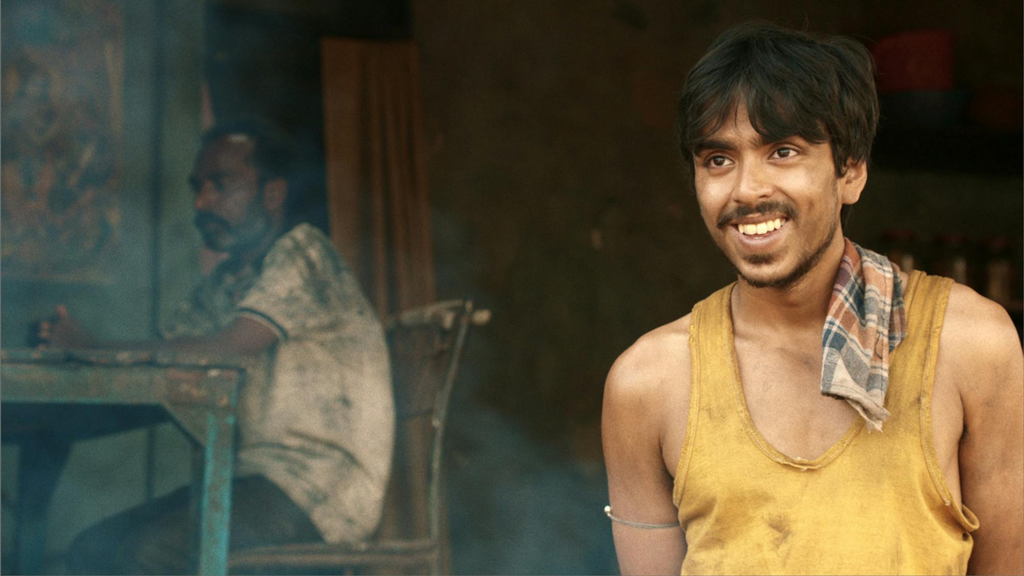 Adarsh Gourav as Balram in a still from the film. He has on a yellow tank top that is dinged with dirt. There’s a blue and red plaid towel laid over his shoulder, and he’s smiling just off camera. 