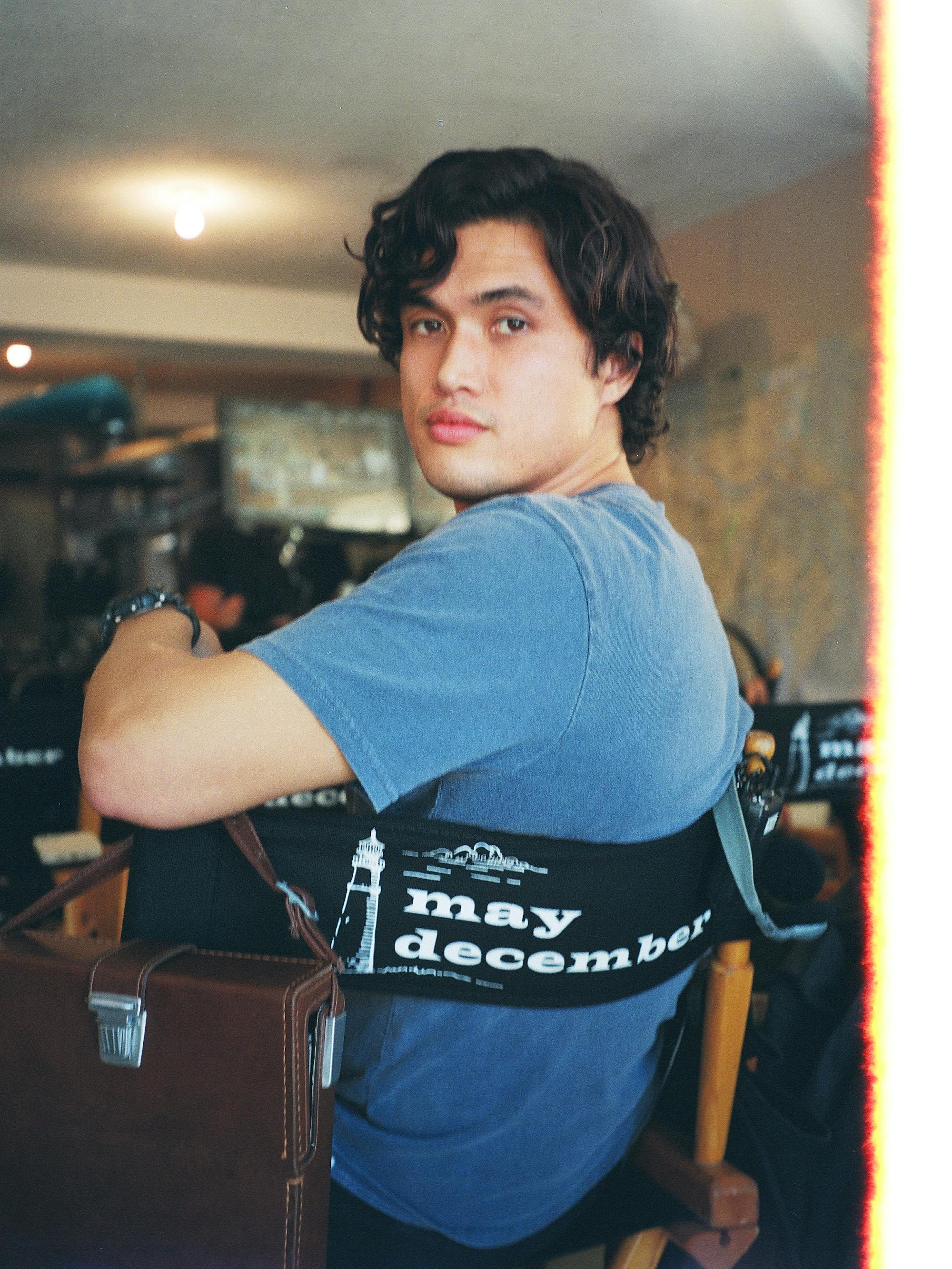 Charles Melton on the set of May December wears a navy shirt and sits in a black chair.