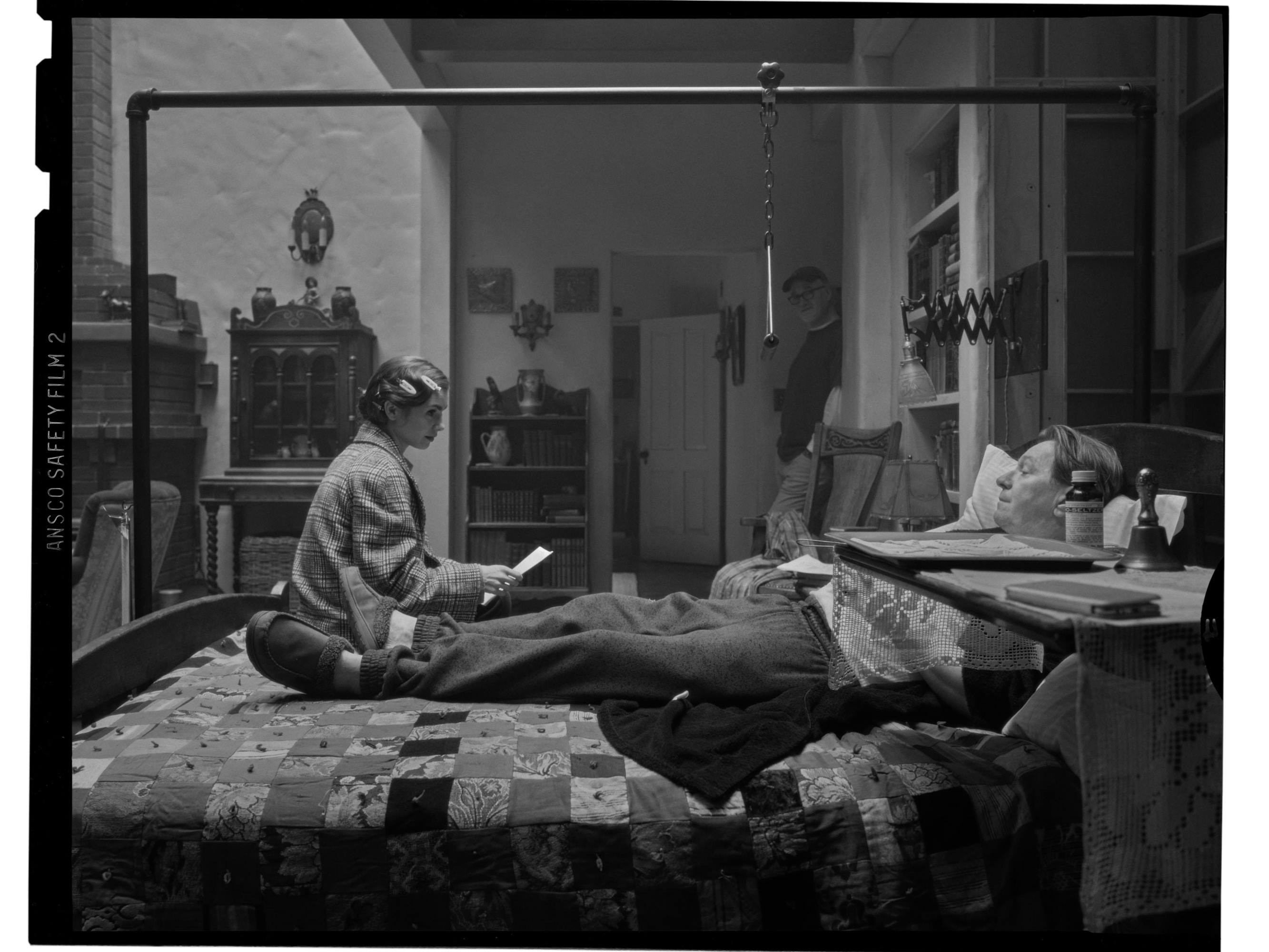 The director and the two actors are shown on a light-filled set. Oldman lies on a bed that’s covered in a quaint quilt. With his head on the pillow, he looks up at Collins, who holds a script in her hands. In the background, Fincher leans against a wall and observes the rehearsal. 
