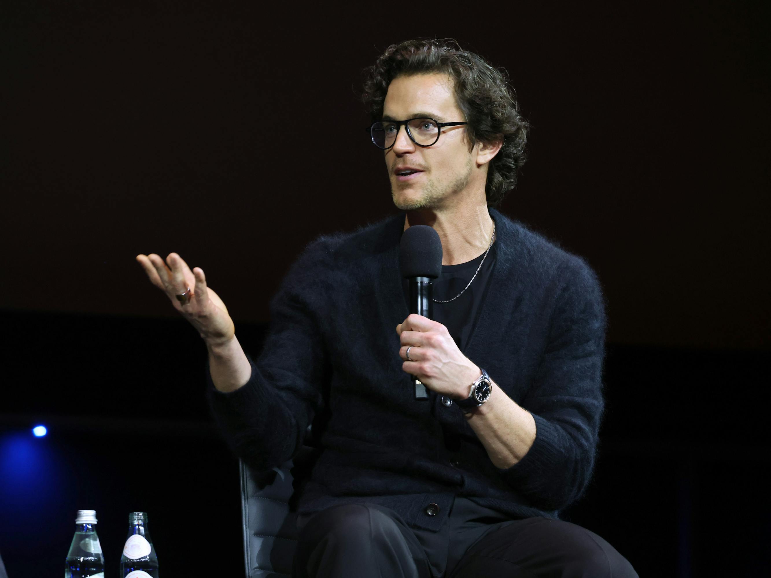 Matt Bomer wears a navy sweater and dark glasses and gestures with his hand.
