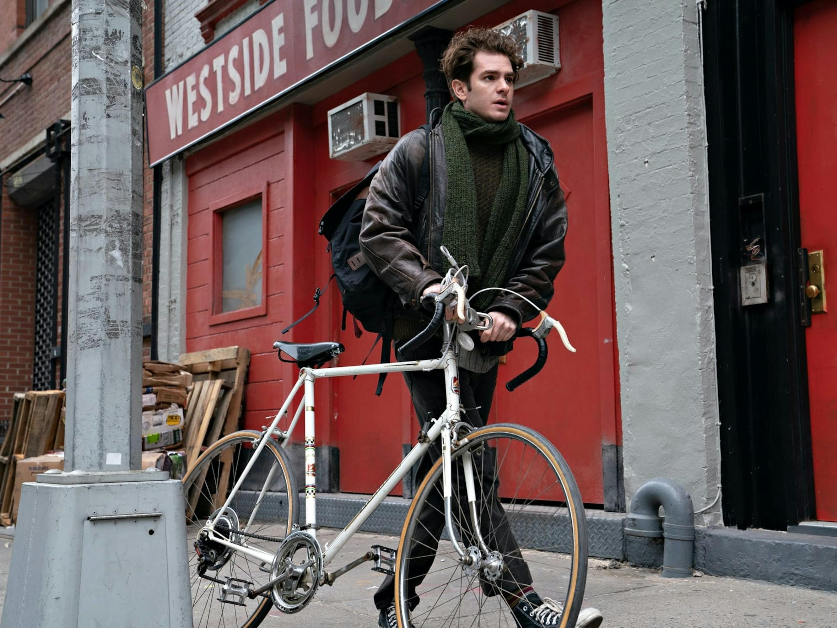 Jonathan Larson wears a brown leather jacket, green scarf, and black converse as he pushes his bike along a New York street.