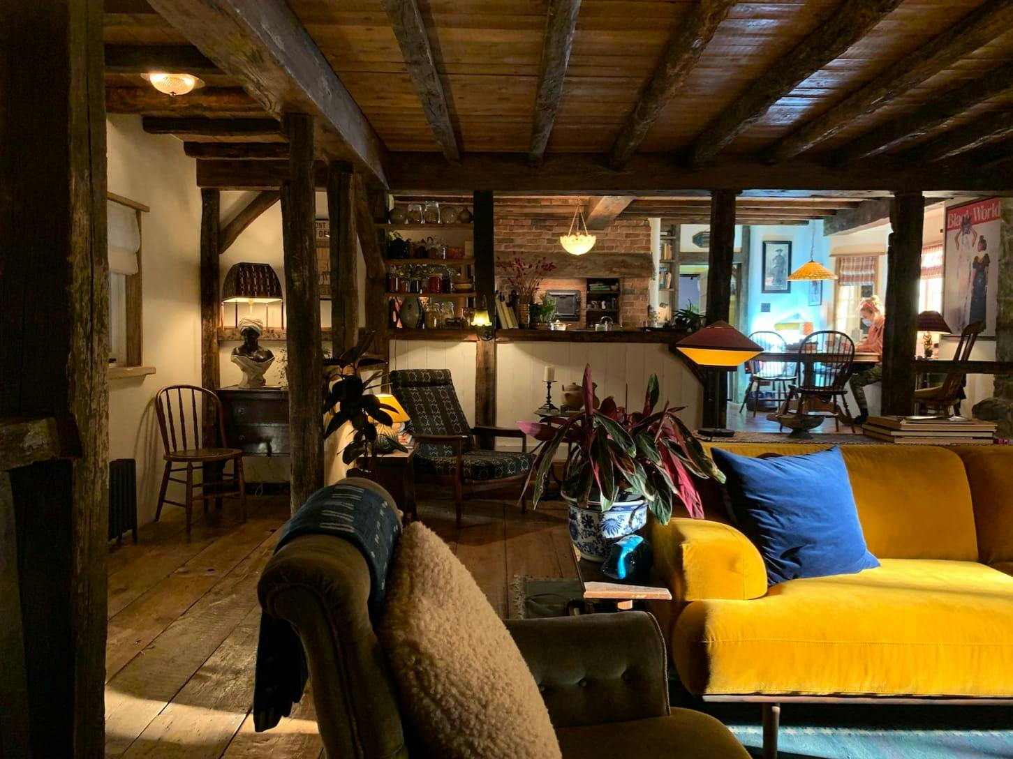 Another shot of the living room complete with yellow couch: brown chairs, wood paneled ceiling, plants, lamps, and a blue pillow.