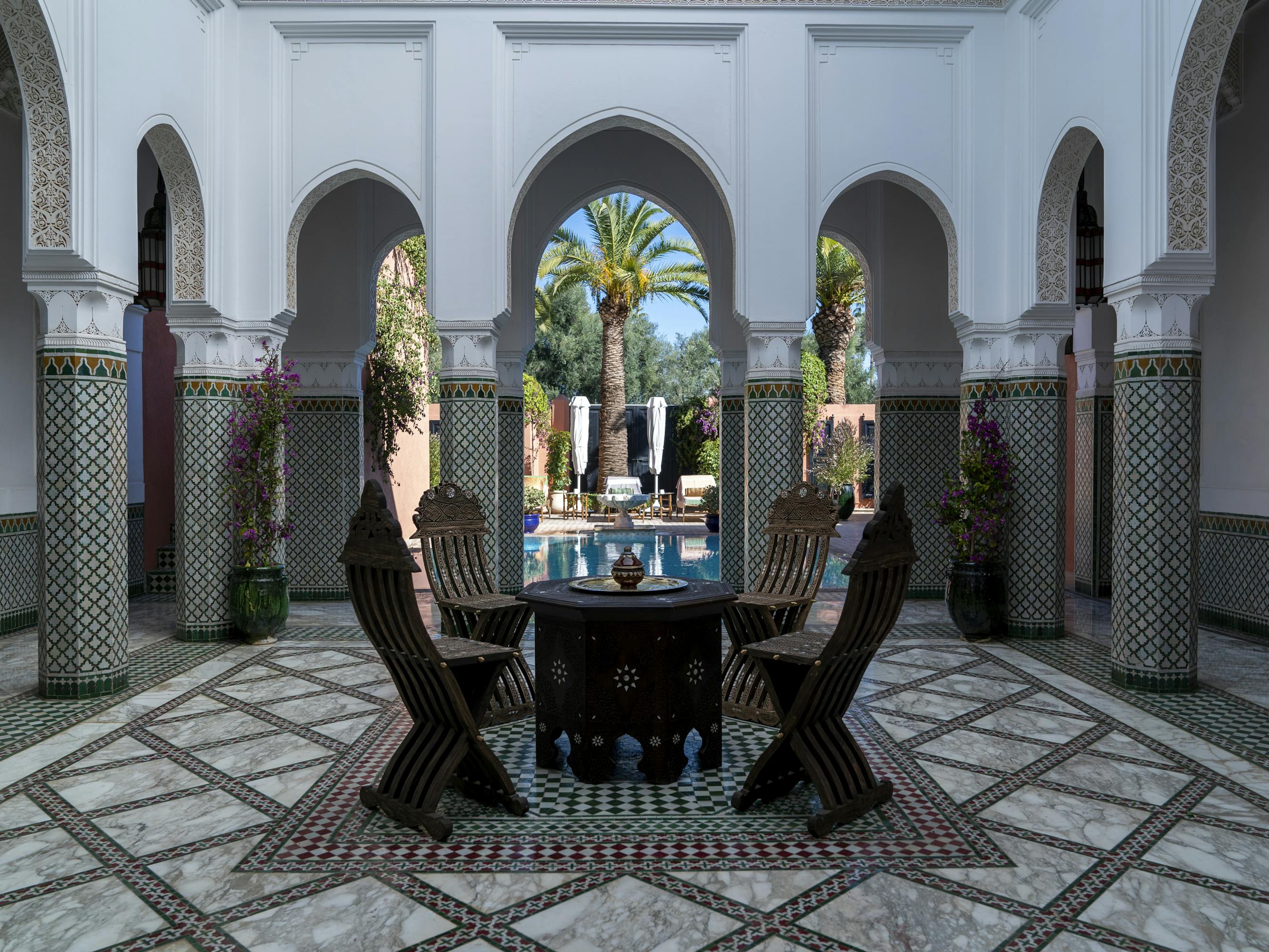 La Mamounia hotel! There is a black table at center, flanked by four chairs. There are pillars surrounding this nook, and a pool can be seen through the pillars.
