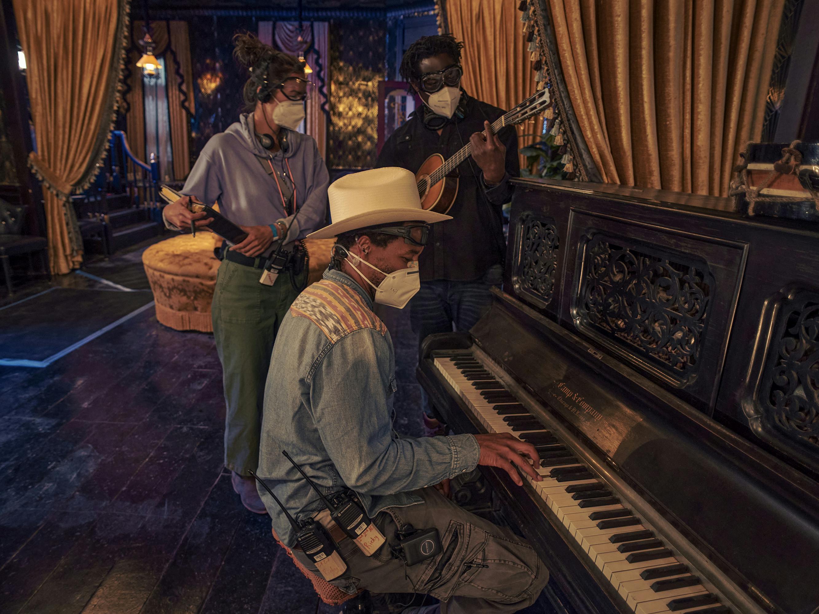 Jeymes Samuel and The Harder They Fall crew stand around a black piano. Behind them are dramatic orange tasseled curtains and dark wooden floors.