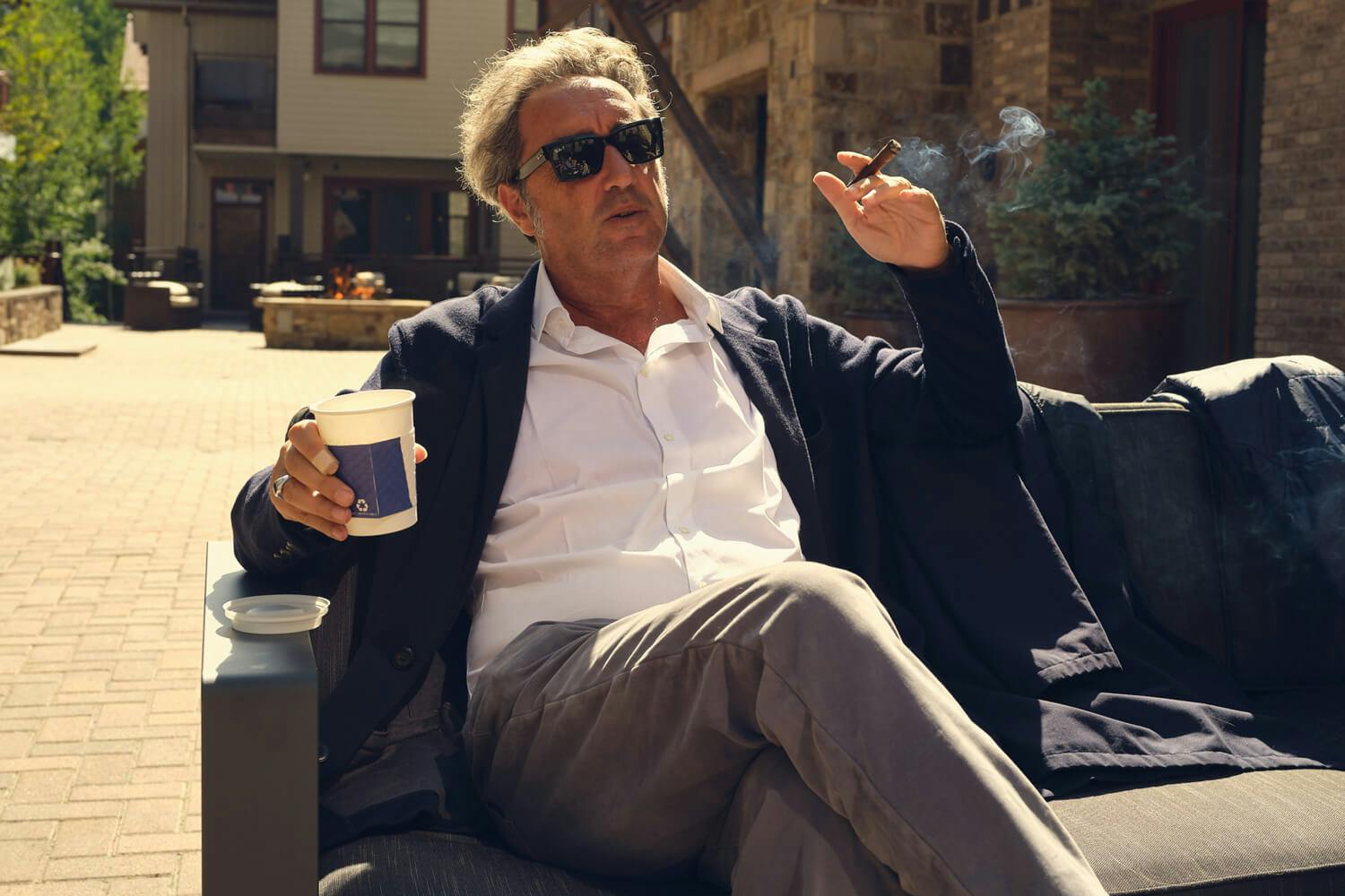 Paolo Sorrentino sits outside at the Telluride Film Festival wearing casual attire while enjoying a cup of coffee and a cigar. 