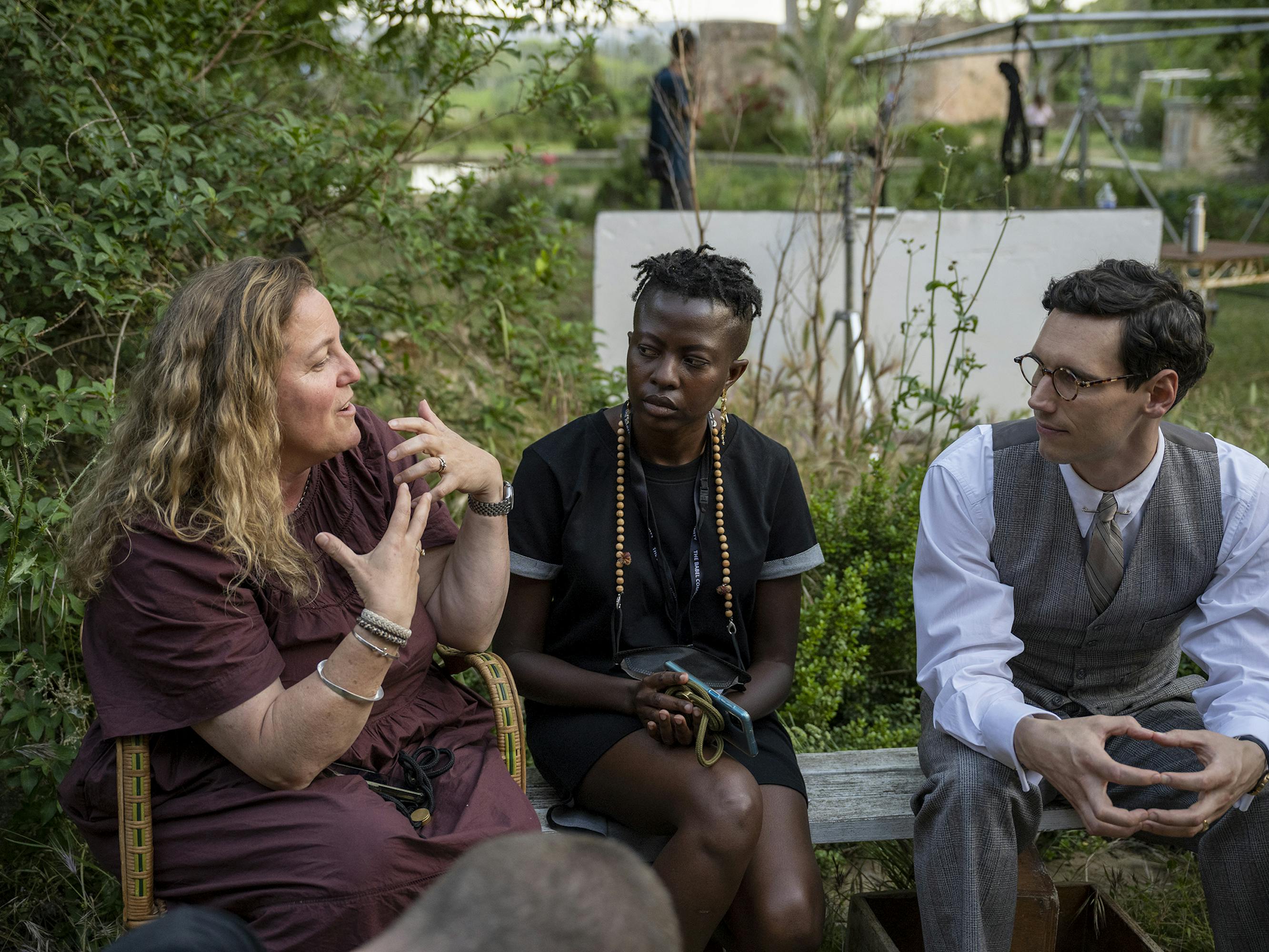 Anna Winger and Cory Michael Smith on the set of Transatlantic. Anna wears a maroon dress, someone sits between them in a black ensemble with tan beads, and Cory wears a three-piece suit vest and pants.