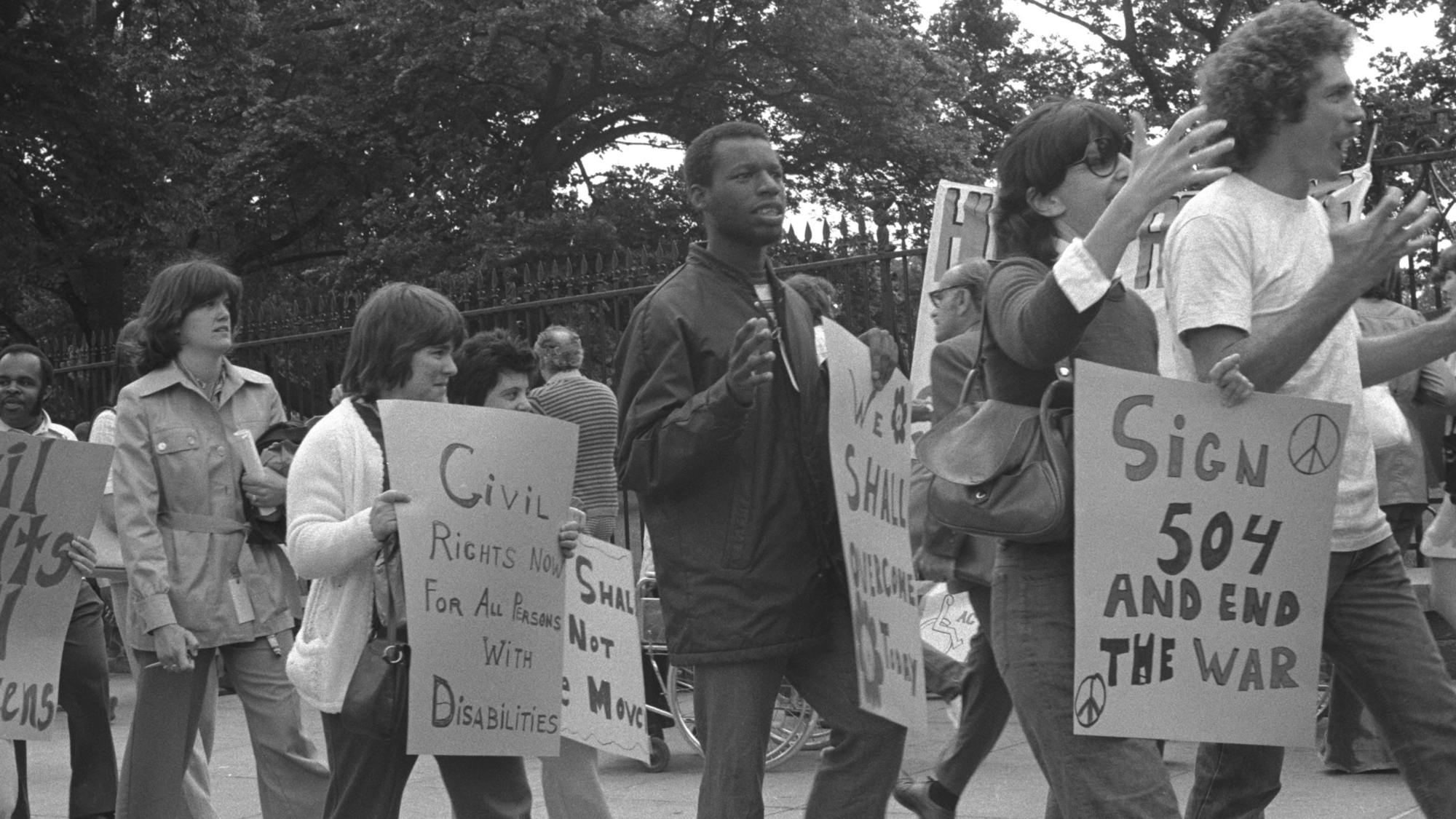 Demonstrators are pictured in black and white, holding signs that read “Civil Rights Now for All Persons with Disabilities” and “Sign 504 and End the War.”
