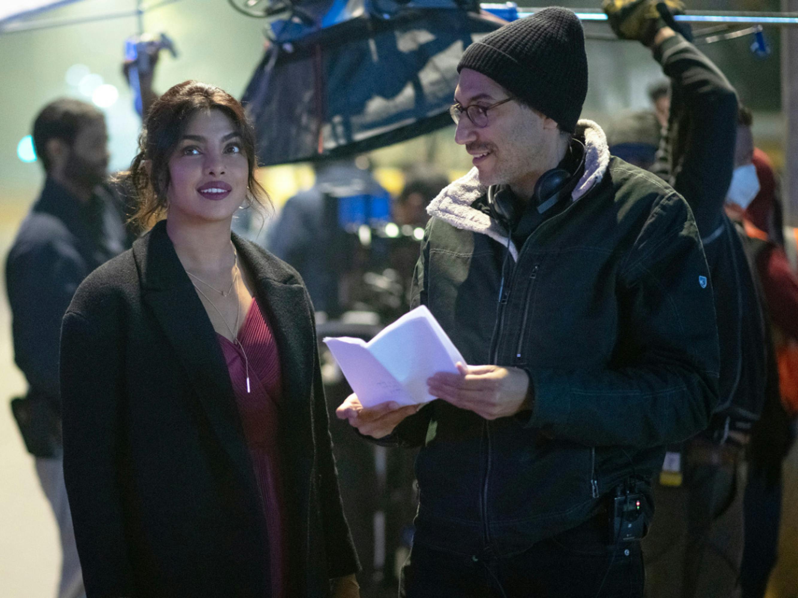 Priyanka Chopra Jonas is seen with director Ramin Bahrani in a behind the scenes photo. In the background, the crew are moving lighting equipment. It looks like it’s nighttime and they are outside. Bahrani is holding some papers, potentially script pages. Both are smiling.