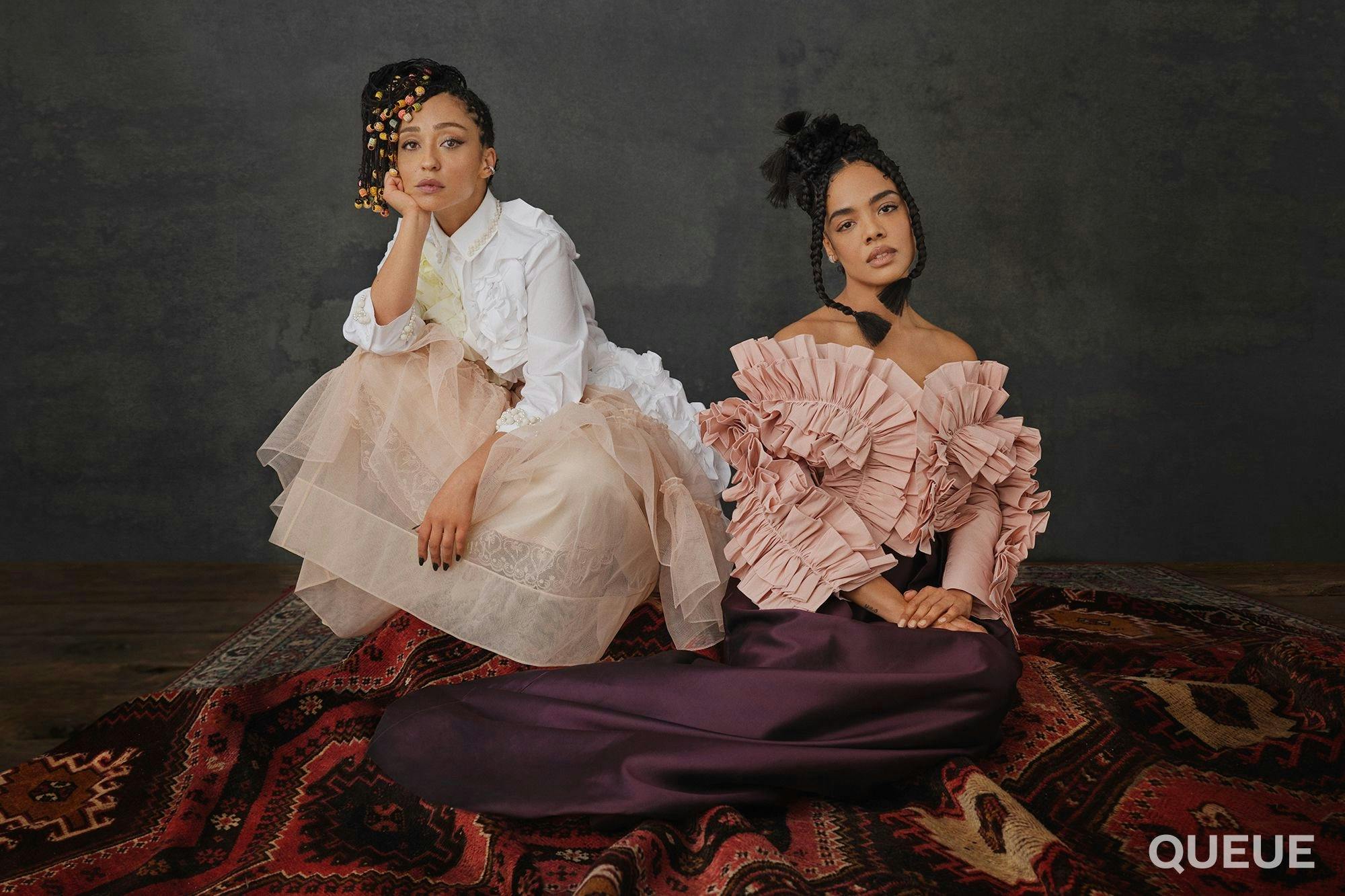 Ruth Negga and Tessa Thompson sit side by side on a pile of colorful rugs.  Ruth wears a peach tulle skirt and a white ruffled shirt.  Tessa wears a long purple skirt and a pale pink ruffled shirt