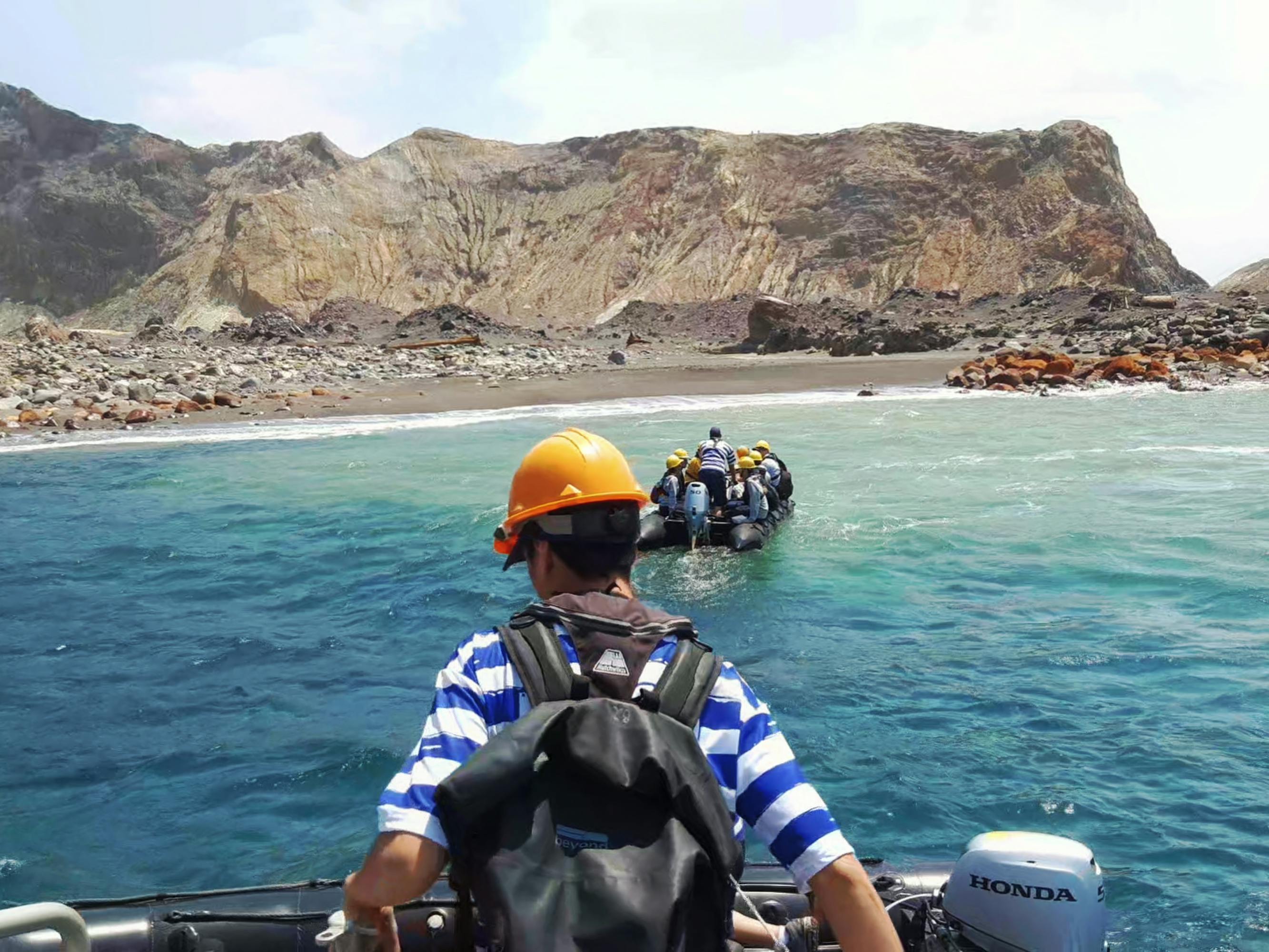 Tourist groups arrive at the volcano in small boats. The man in the foreground wears a white and blue striped shirt, and a yellow hard hat. 