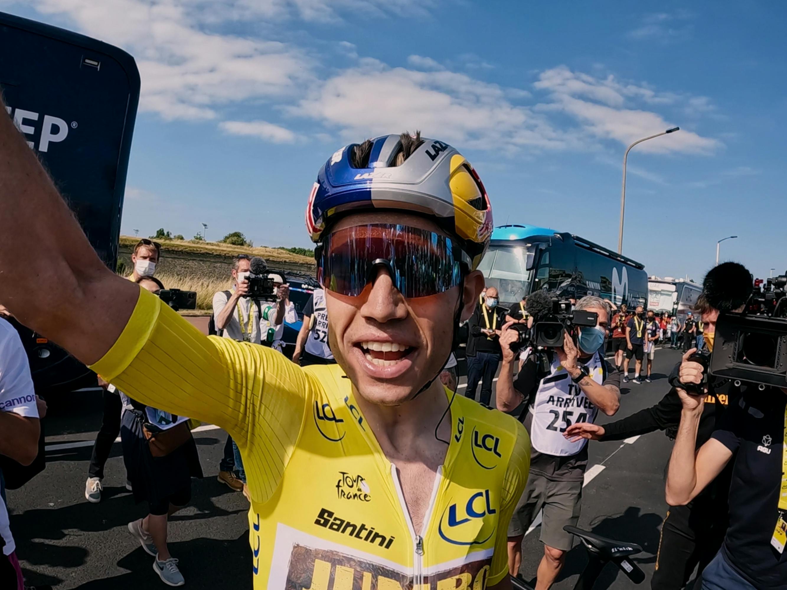 A cyclist wears the yellow jersey and smiles, with one arm raised.